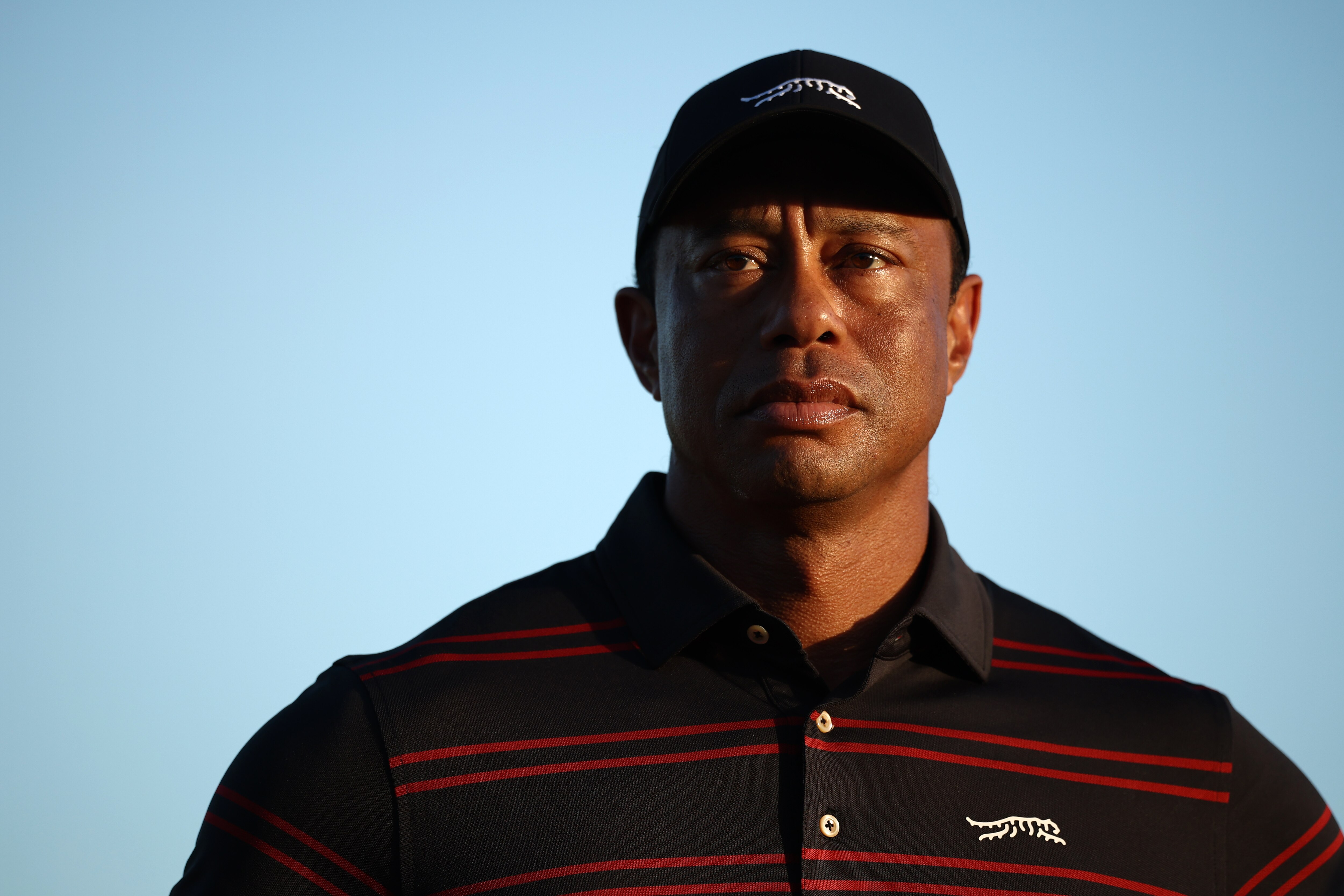 Close-up portrait of a Tiger Woods wearing a black baseball cap and a black collared shirt with thin red stripes, standing against a clear blue sky. Warm, low-angle sunlight illuminates one side of his face, casting soft shadows as he looks ahead with a serious, focused expression.