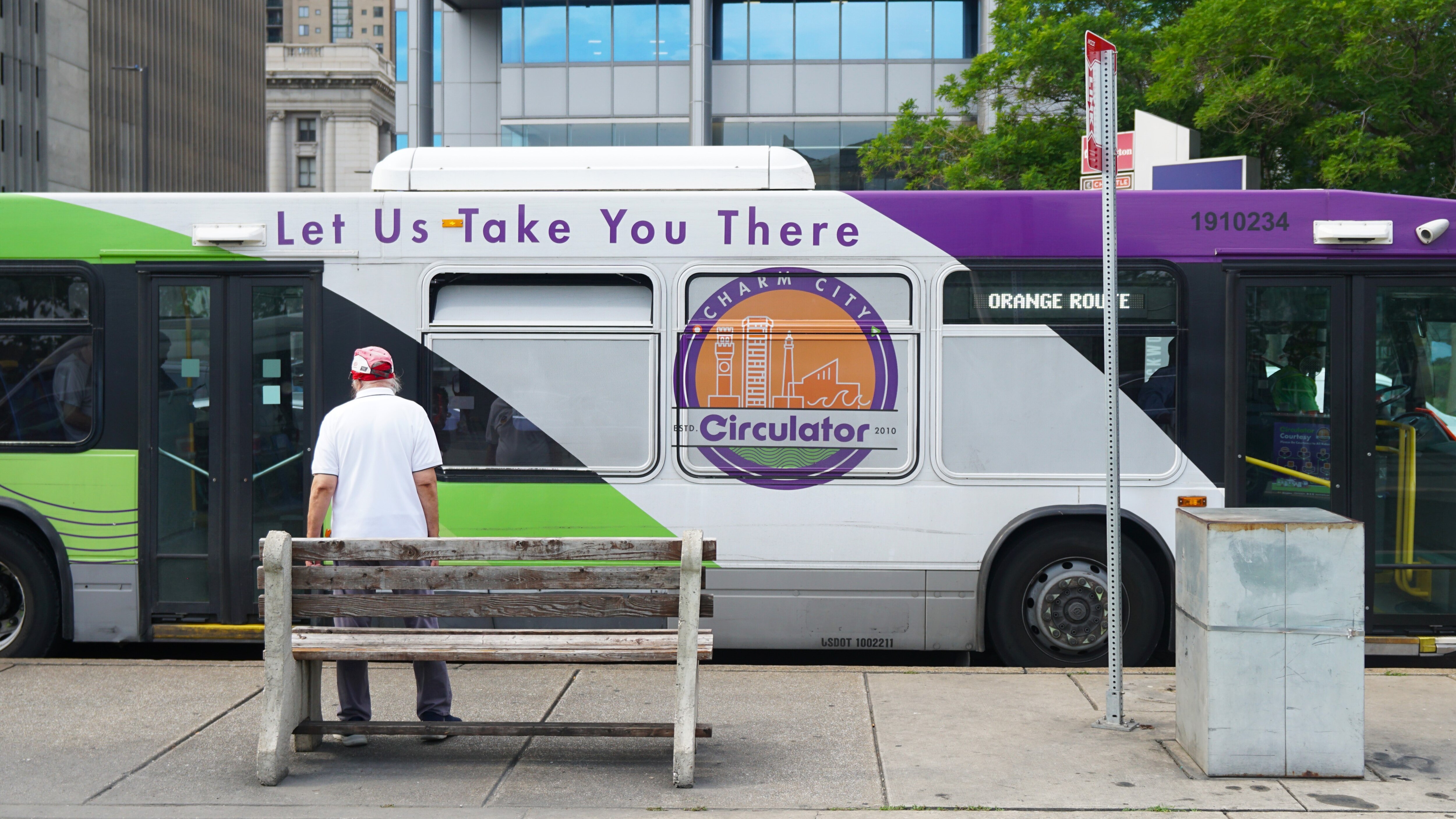 A green, white and purple bus stops at a bus stop in front of a man standing by a bench.