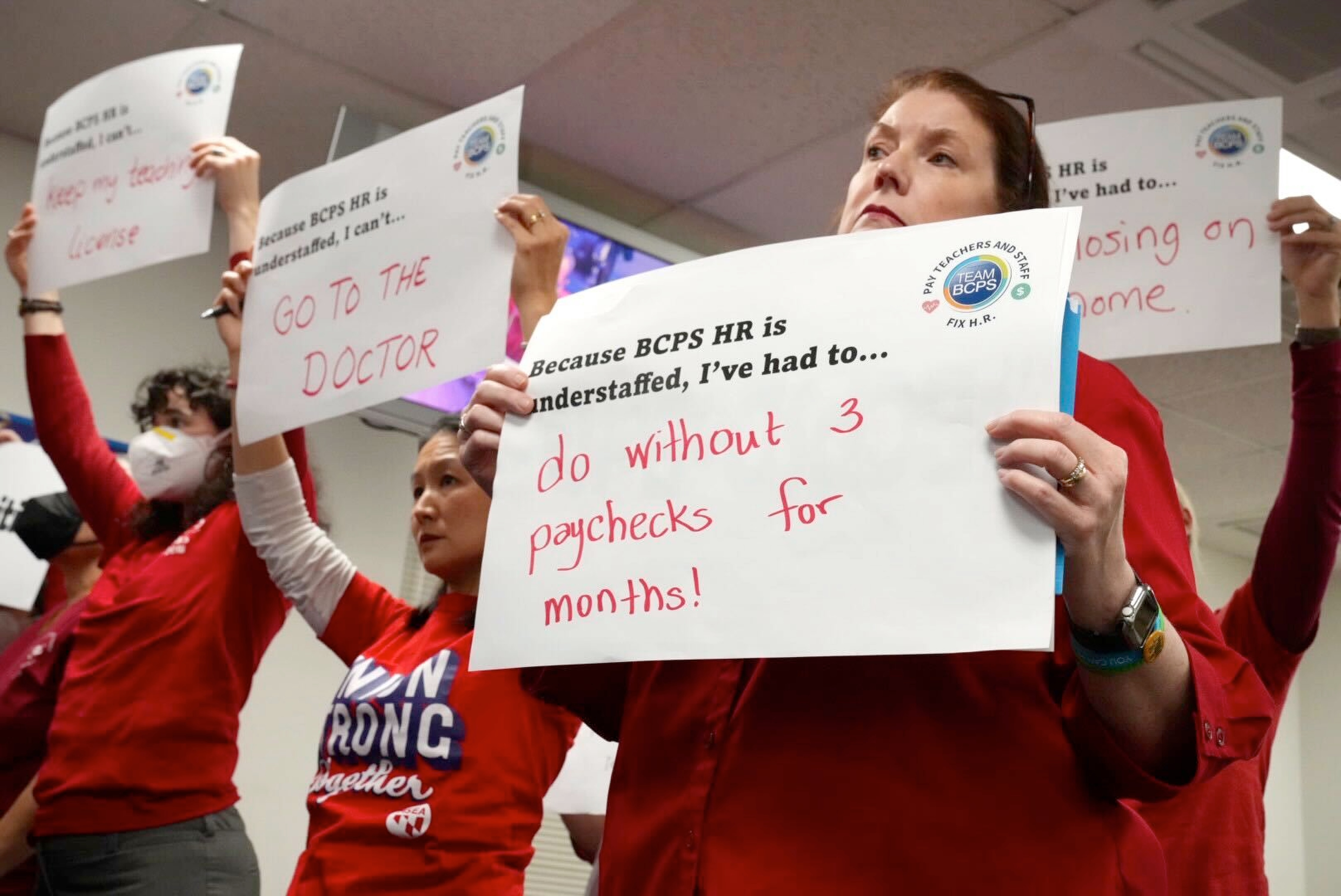 Kathleen Cave, right, and other Baltimore County Public School teachers stand together with signs calling out BCPS for pay being withheld, mistakes with benefits, delays in tuition reimbursement and stalled certification, during a school board meeting Tuesday, December 6.