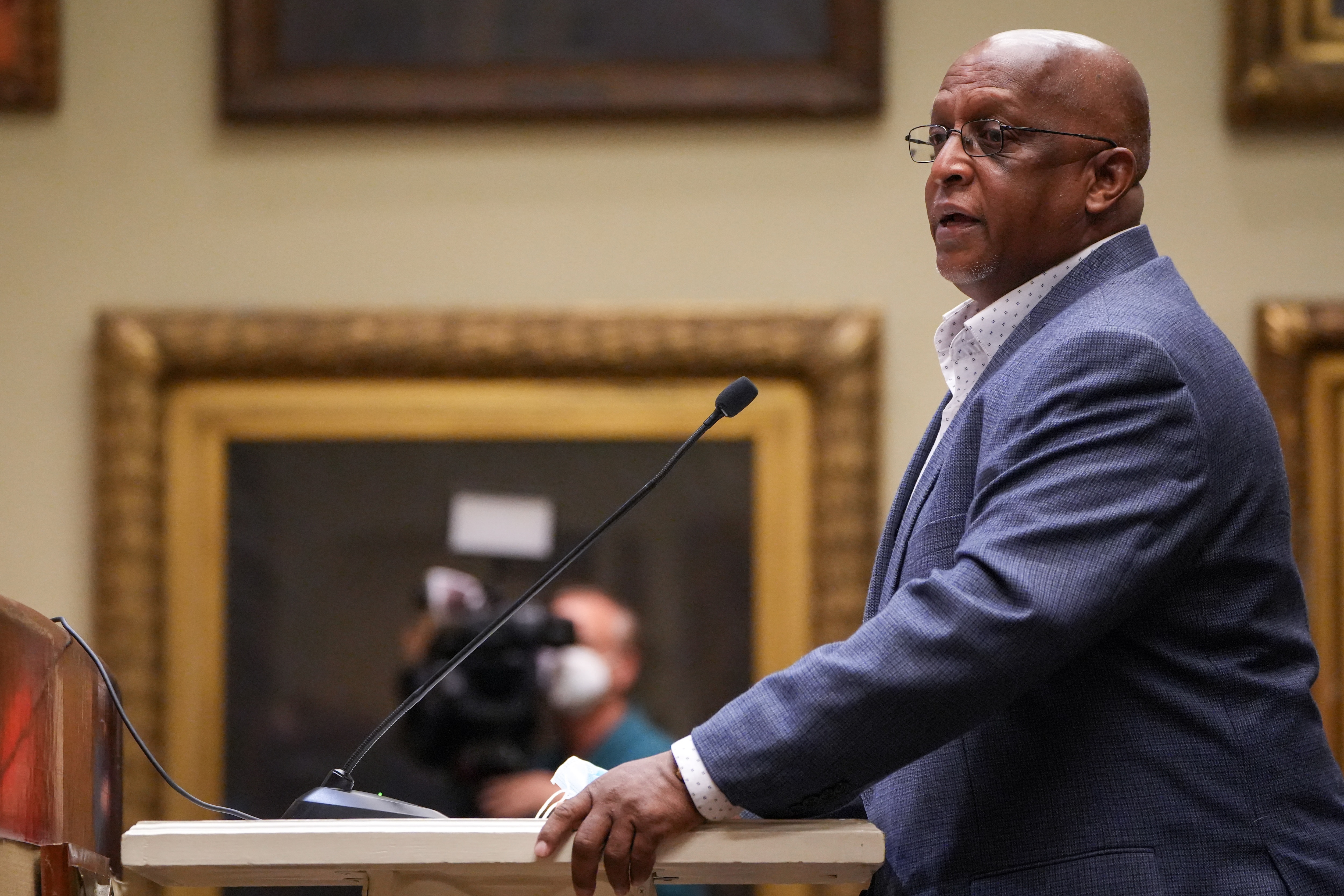 Former Baltimore Mayor Bernard C. “Jack” Young speaks at a lectern during a Baltimore City Board of Estimates meeting inside City Hall. He wears a blue blazer and a white dress shirt.