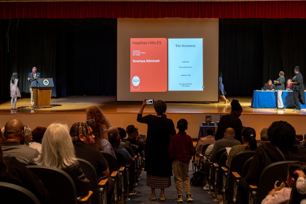 Soumya Alimineti, 11, left, is introduced on stage while her mother Hima Jandhyala, center, films video during the award ceremony for the Prince George's County Write-A-Book contest.