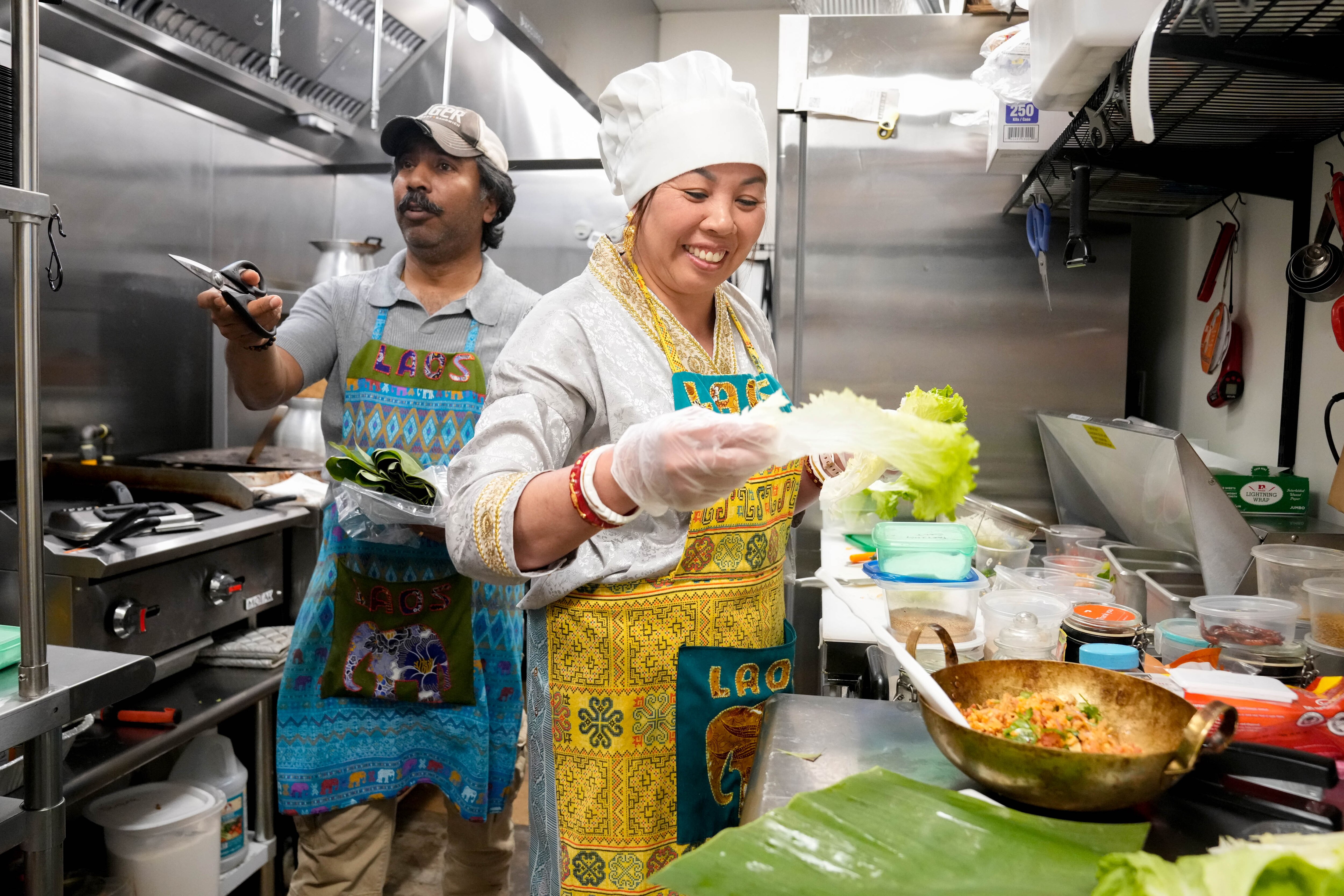 Saroj Sharma, left, manager at Baltimore Lao Eats, explains the dish that his wife, Khamhou Thepsouvanh, the restaurant’s owner and chef, is preparing in their kitchen in Baltimore.