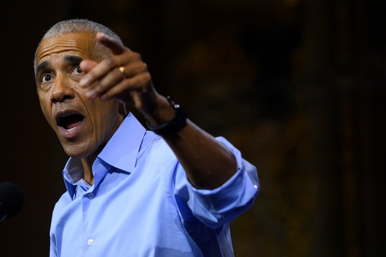 Barack Obama wears a blue shirt with his sleeves rolled up. His mouth is open as he speaks and his left arm is raised and pointing.