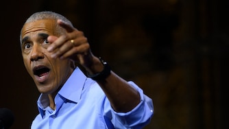 Barack Obama wears a blue shirt with his sleeves rolled up. His mouth is open as he speaks and his left arm is raised and pointing.
