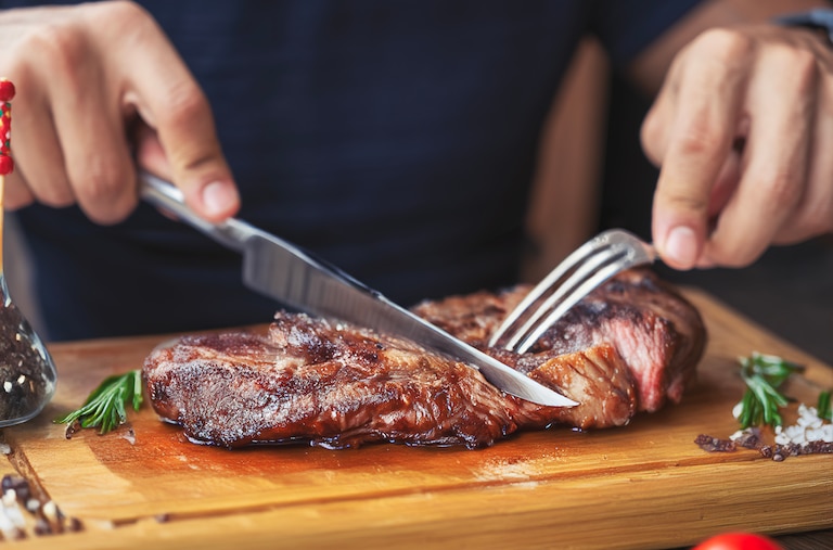 Hands holding fork and knife and eating delicious juicy steak on a wooden plate.