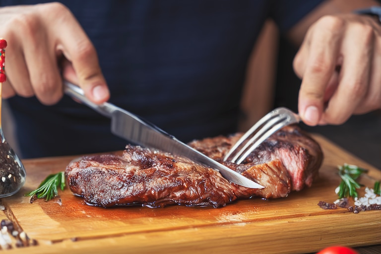 Hands holding fork and knife and eating delicious juicy steak on a wooden plate.