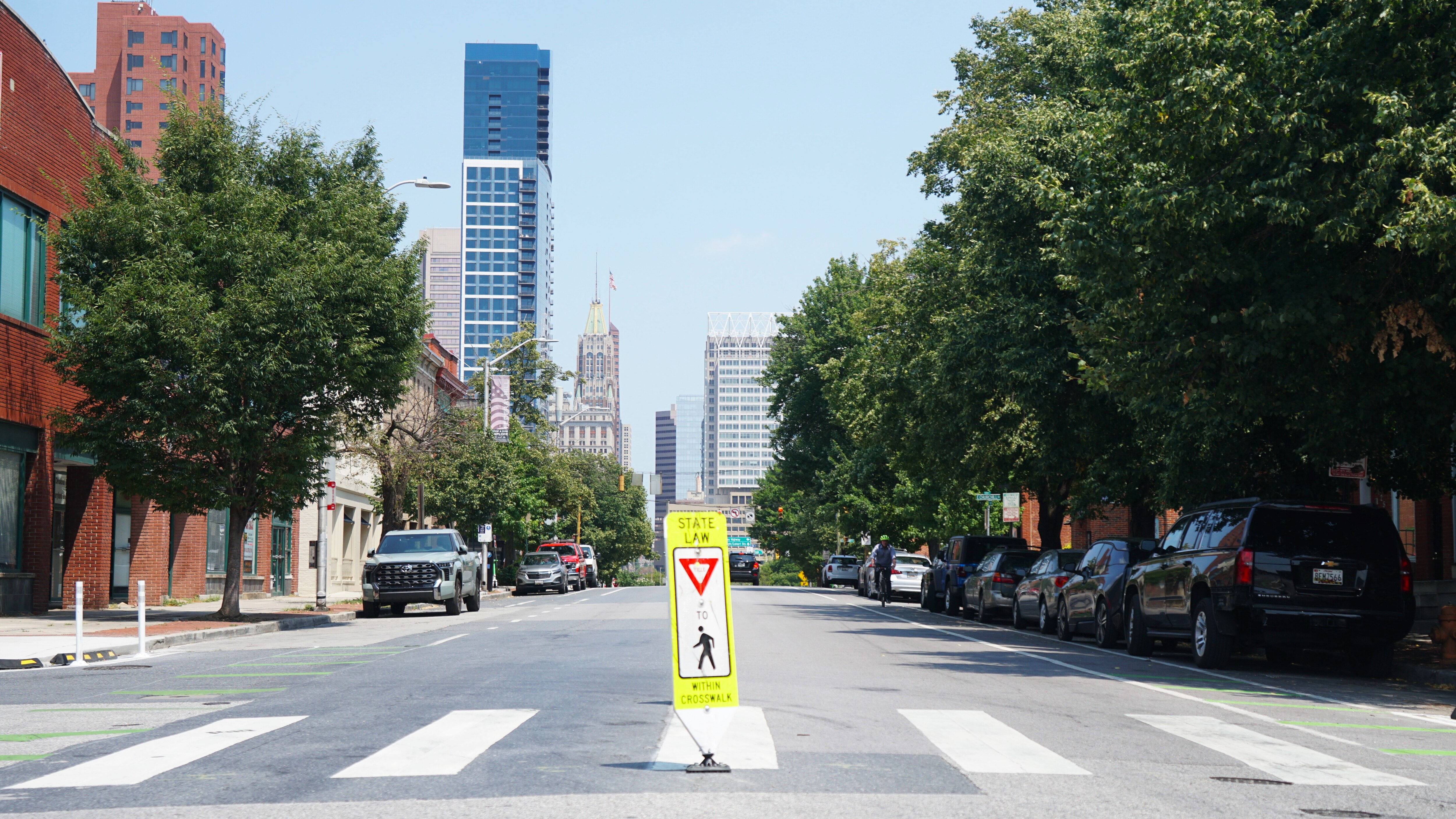 A yellow sign, warning cars about pedestrians, is shown in the middle of a roadway intersection. Cars and trees line the road and a couple of large skyscrapers are in the background.