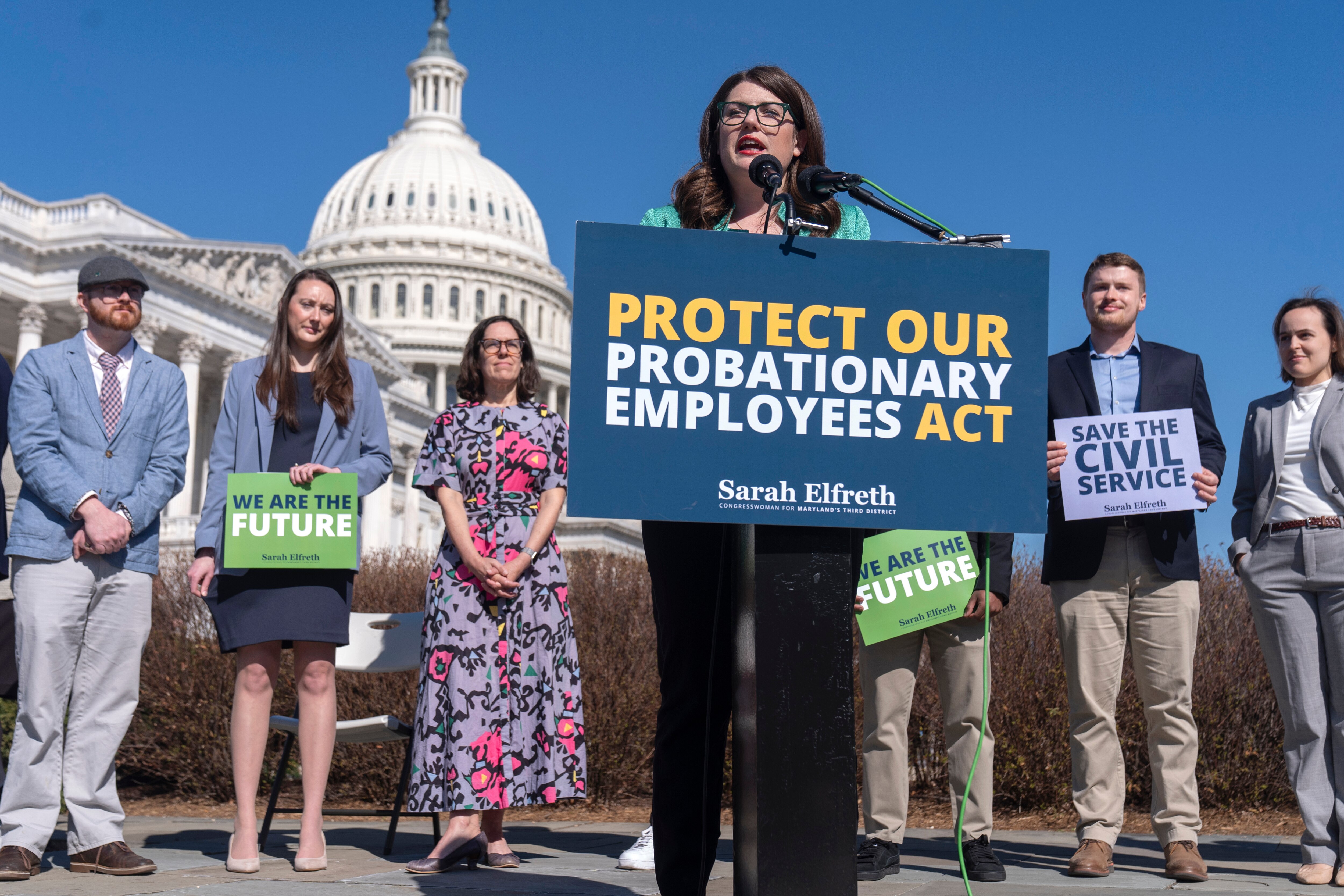 Rep. Sarah Elfreth stands behind a sign that says "PROTECT OUR PROBATIONARY EMPLOYEES ACT." Fired probationary employees stand behind her and the Capitol is in the background.