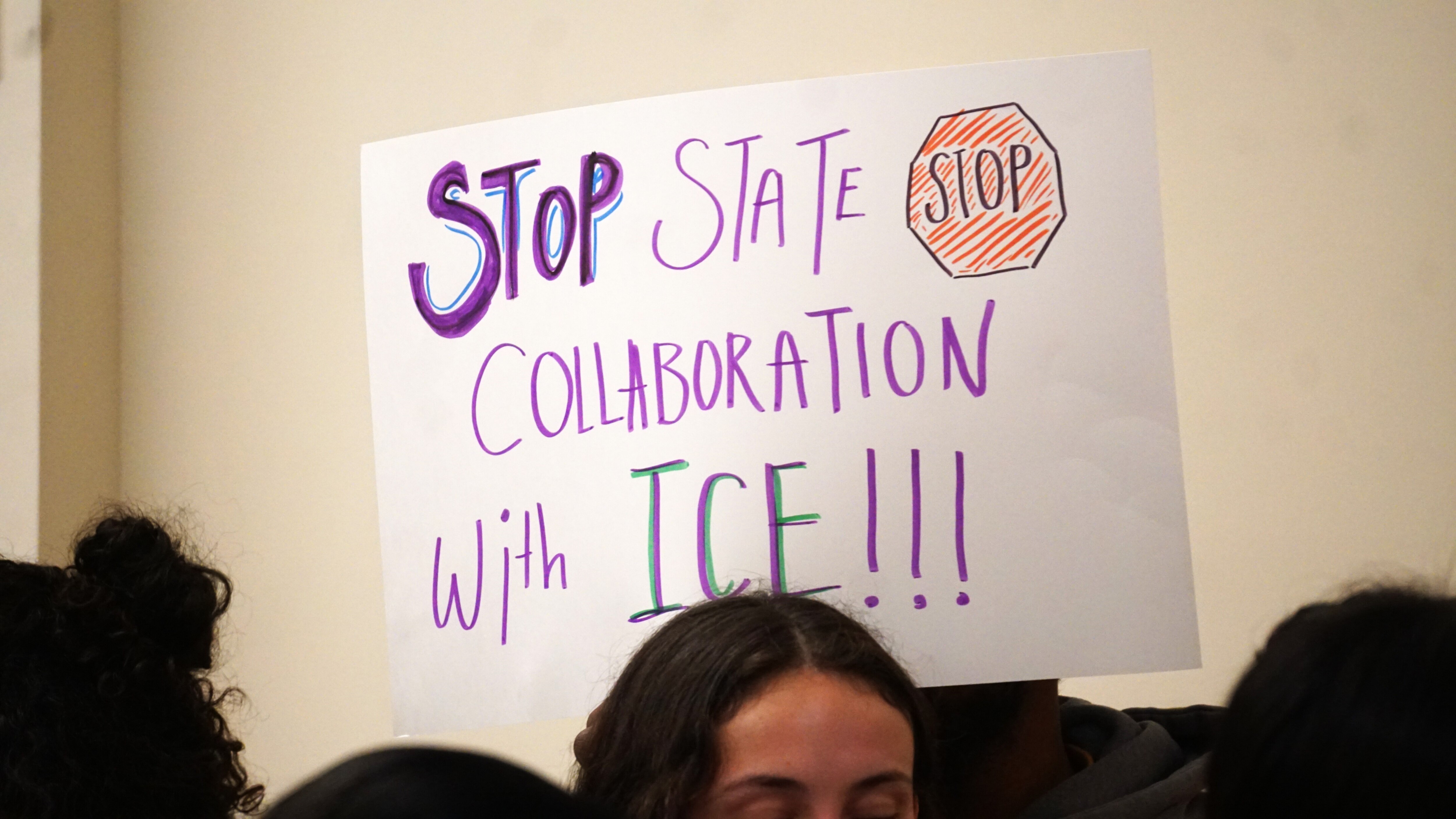 A white poster board sign that reads "STOP STATE COLLABORATION WITH ICE!!!"