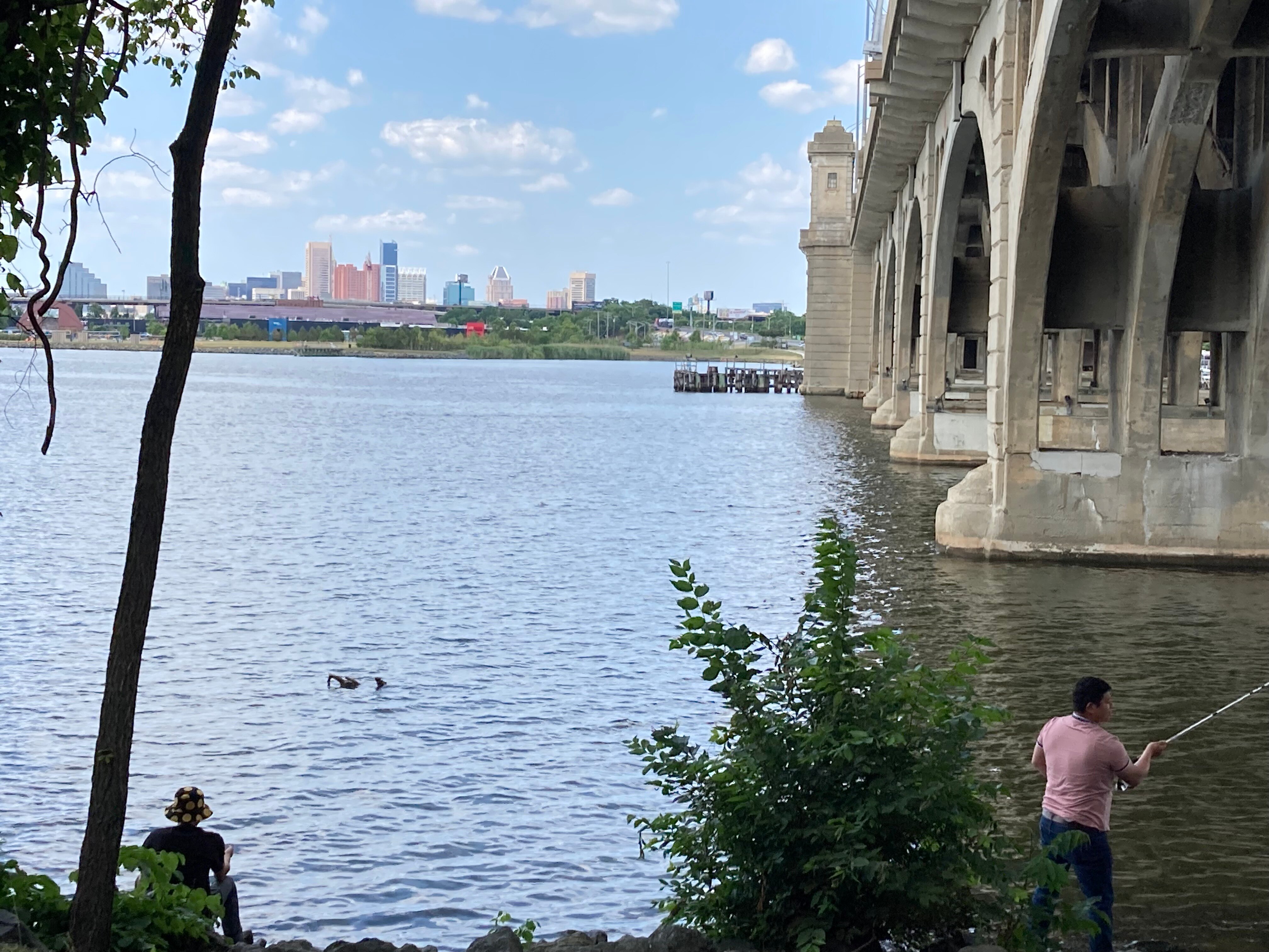 Men fish beneath the Hanover Street Bridge in the Middle Branch of the Patapsco River. The downtown Baltimore skyline is visible in the distance. June 4, 2023.