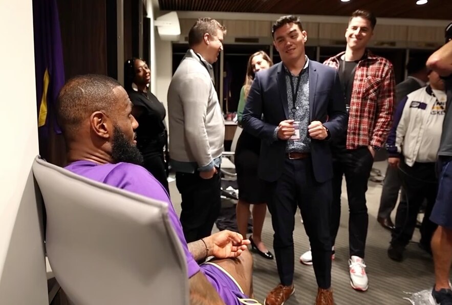 Kyle Goon, The Baltimore Banner's new sports columnist, telling LeBron James that he's leaving California to return to Baltimore. Goon is standing and wearing a suit jacket, smirking. James is seated and we can only see the side of his face.
