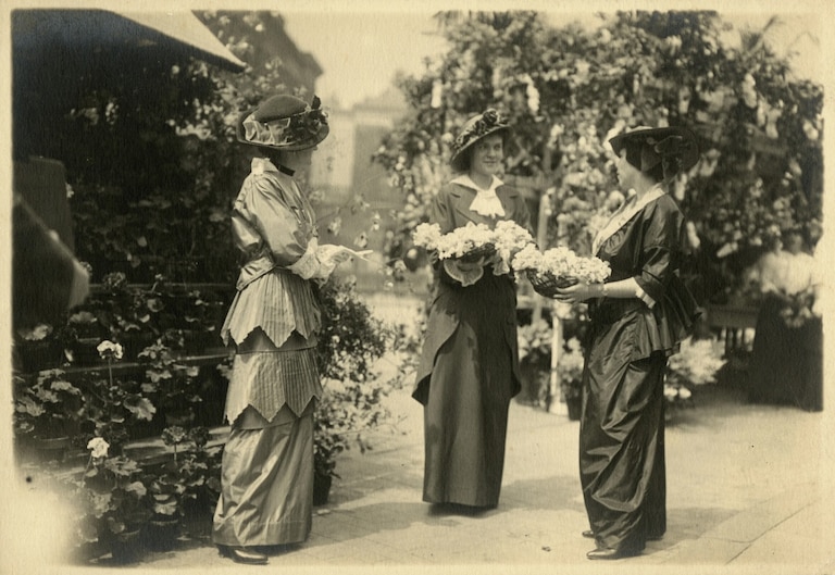 Three individuals at the Flower Mart in the Mount Vernon neighborhood of Baltimore, Maryland. Two carry flowers in their arms. Founded in 1911 by the Women's Civic League, the Flower Mart is the oldest free public festival in Baltimore.