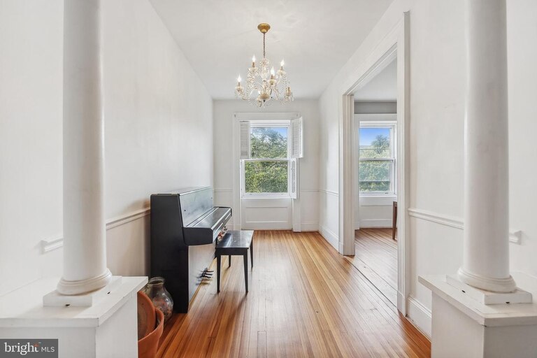 A photo from a real estate listing shows a black piano and piano bench positioned against the wall of a bright room painted white, with two columns in the foreground, a chandelier overhead and hardwood floors that are stained in warm tones. Two windows appear on the far wall and frame trees and blue sky.