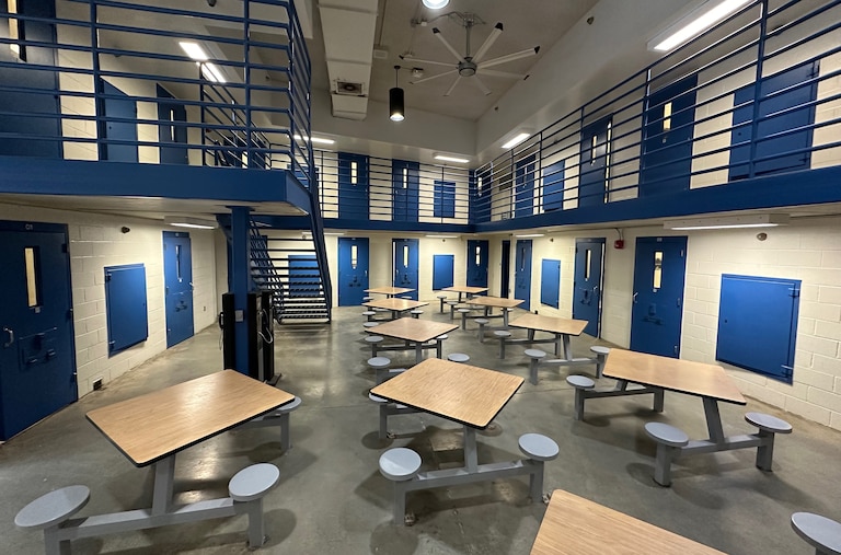 January 22, 2024 - Photo of common area with tables and stools inside prison. Blue doors lead to cells off the ground floor and off a second floor walkway above.