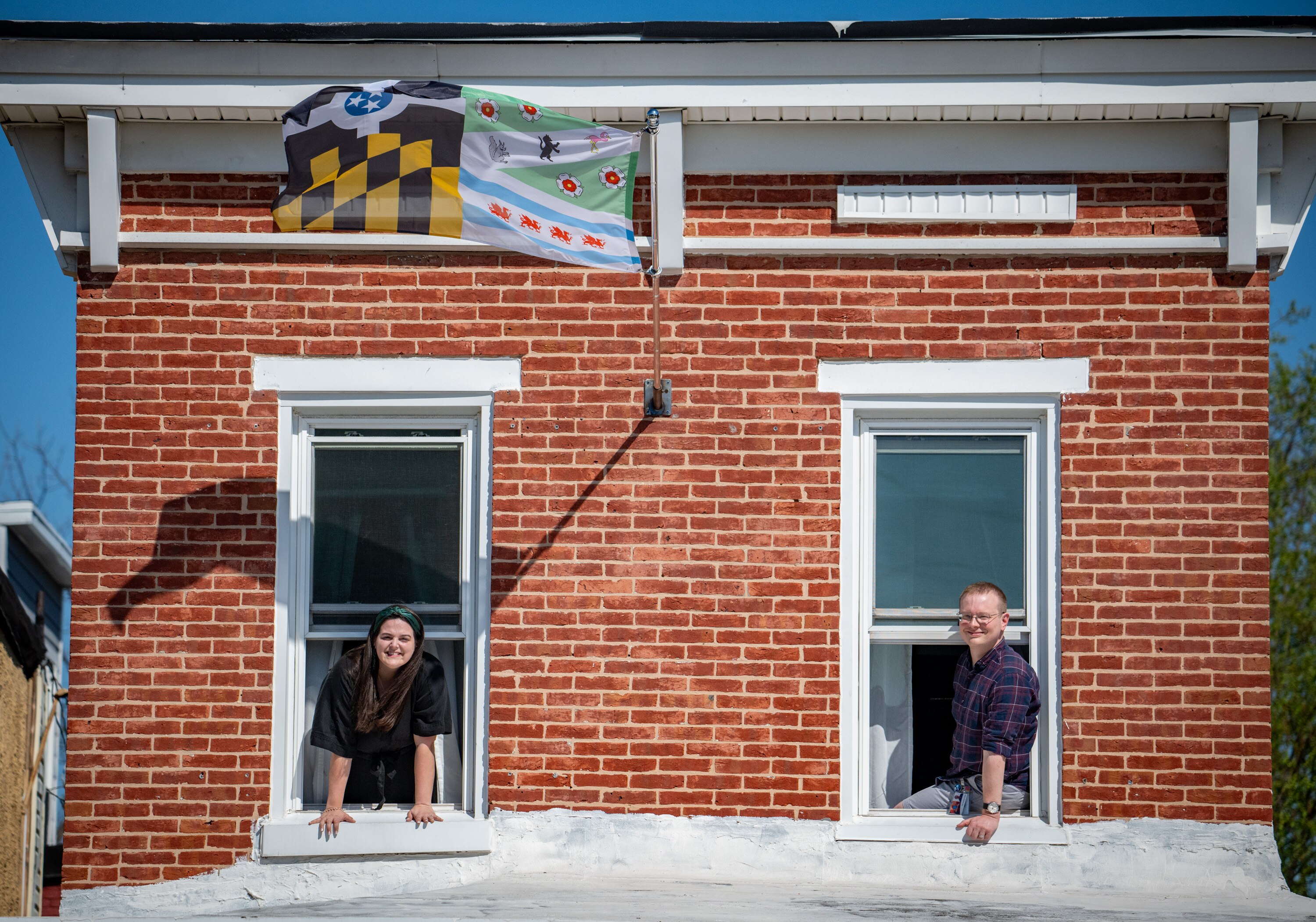 Emily Davidson and Christopher Clark hang out their second story windows below a flag they designed for their upcoming wedding. The couple has flown a variety of flags above their Hampden home. 