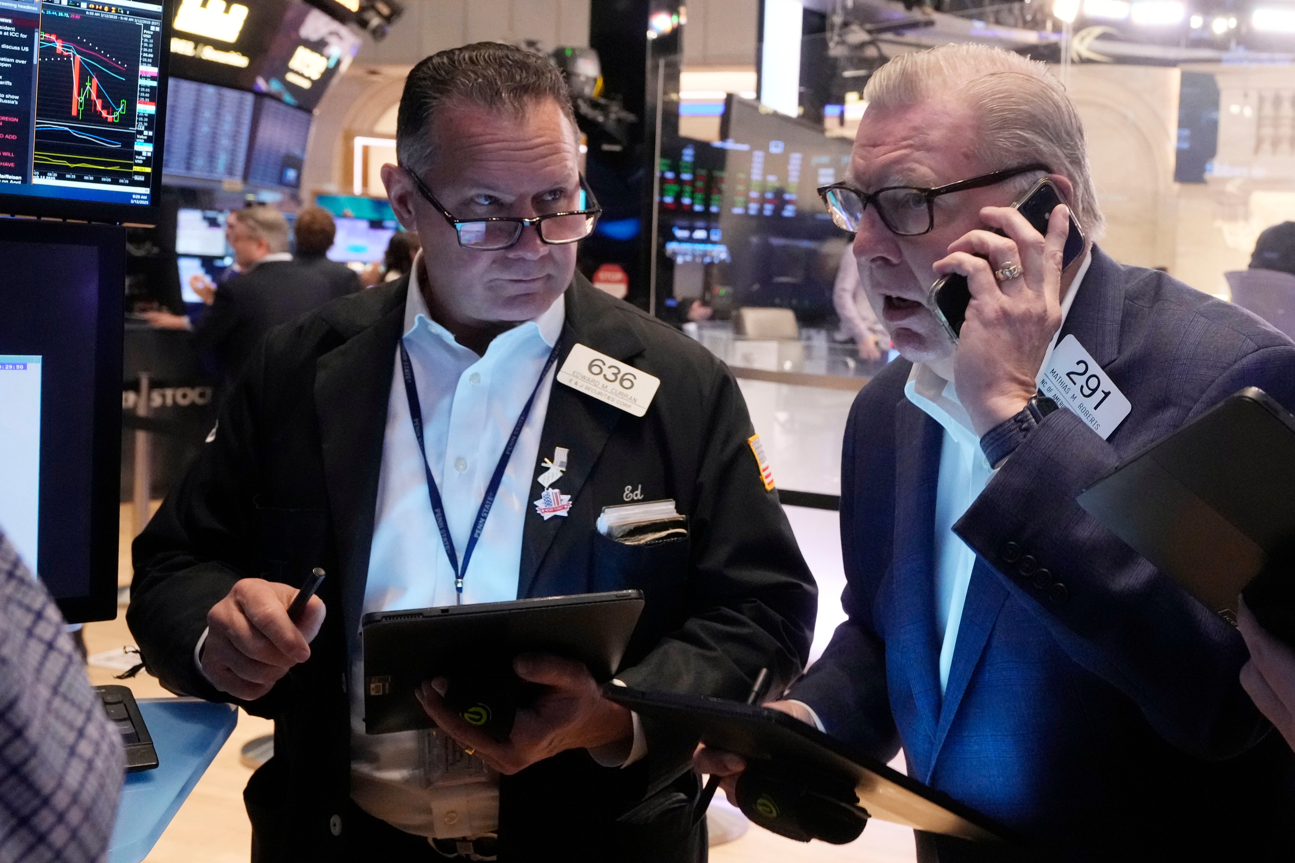 Two male stock traders, both holding tablets and wearing glasses, are see on the floor of the New York Stock Exchange. The one on the right is speaking into a cellphone while the one on the left looks on.