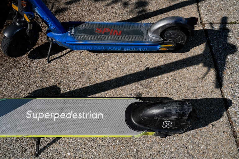 Parked Spin and Superpedestrian scooters are photographed from above on a sidewalk.