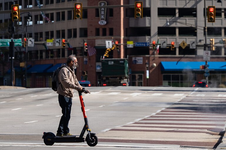 A man scooters across Pratt Street, which is mostly empty behind him and rows of red traffic lights hang above.