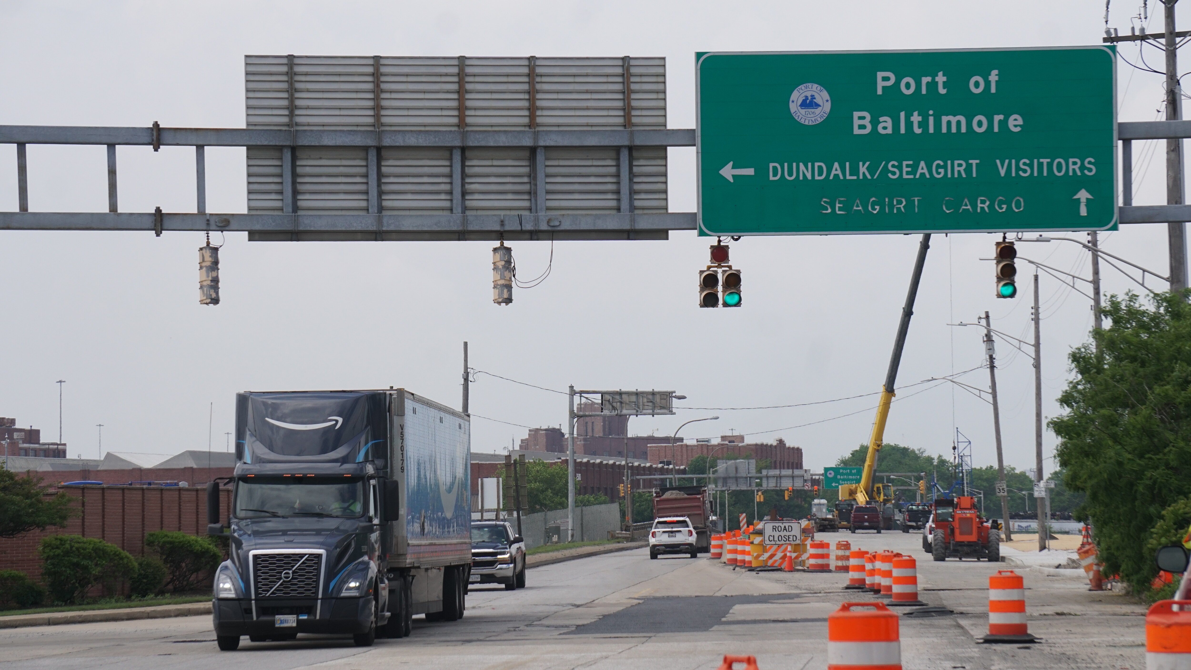 This photo shows a roadway partially closed down with orange construction cones. A truck is driving toward the camera underneath street signs indicating where to turn into a Port of Baltimore terminal.