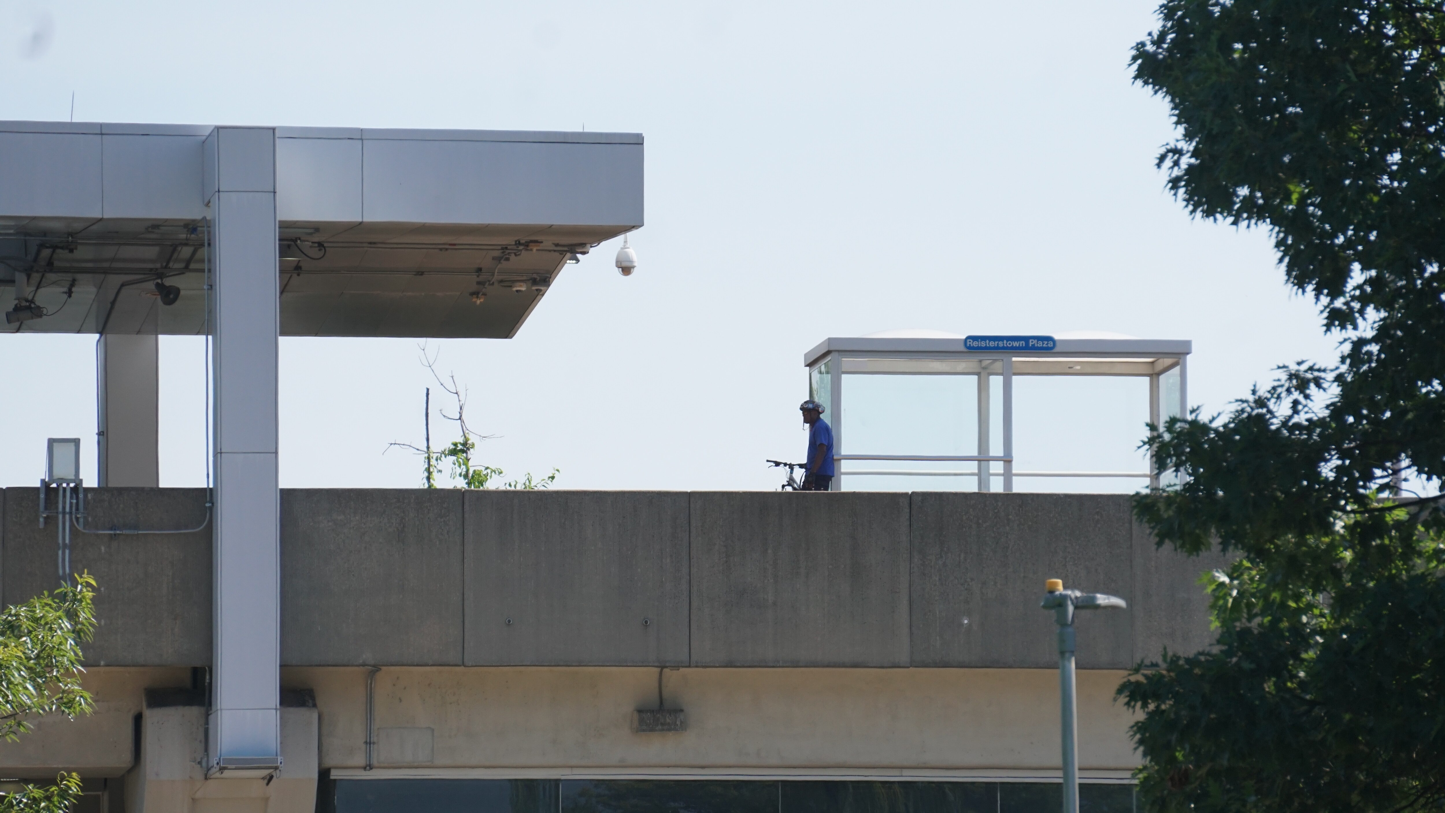 A man on a bike wearing a helmet waits outside of a glass shelter at an above ground train station.