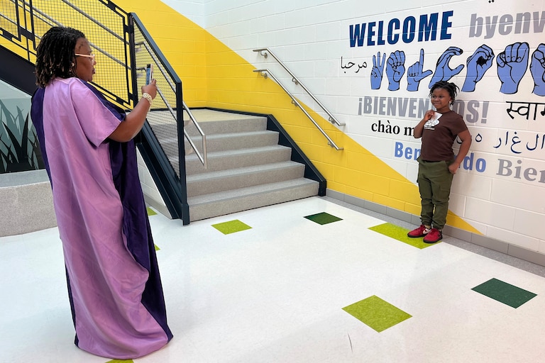 Alica Shaw snaps a picture of her 8-year-old daughter Melvina in front of a mural welcoming students in a dozen languages. Melvina is holding a piece of the ribbon that was cut in the opening ceremony.