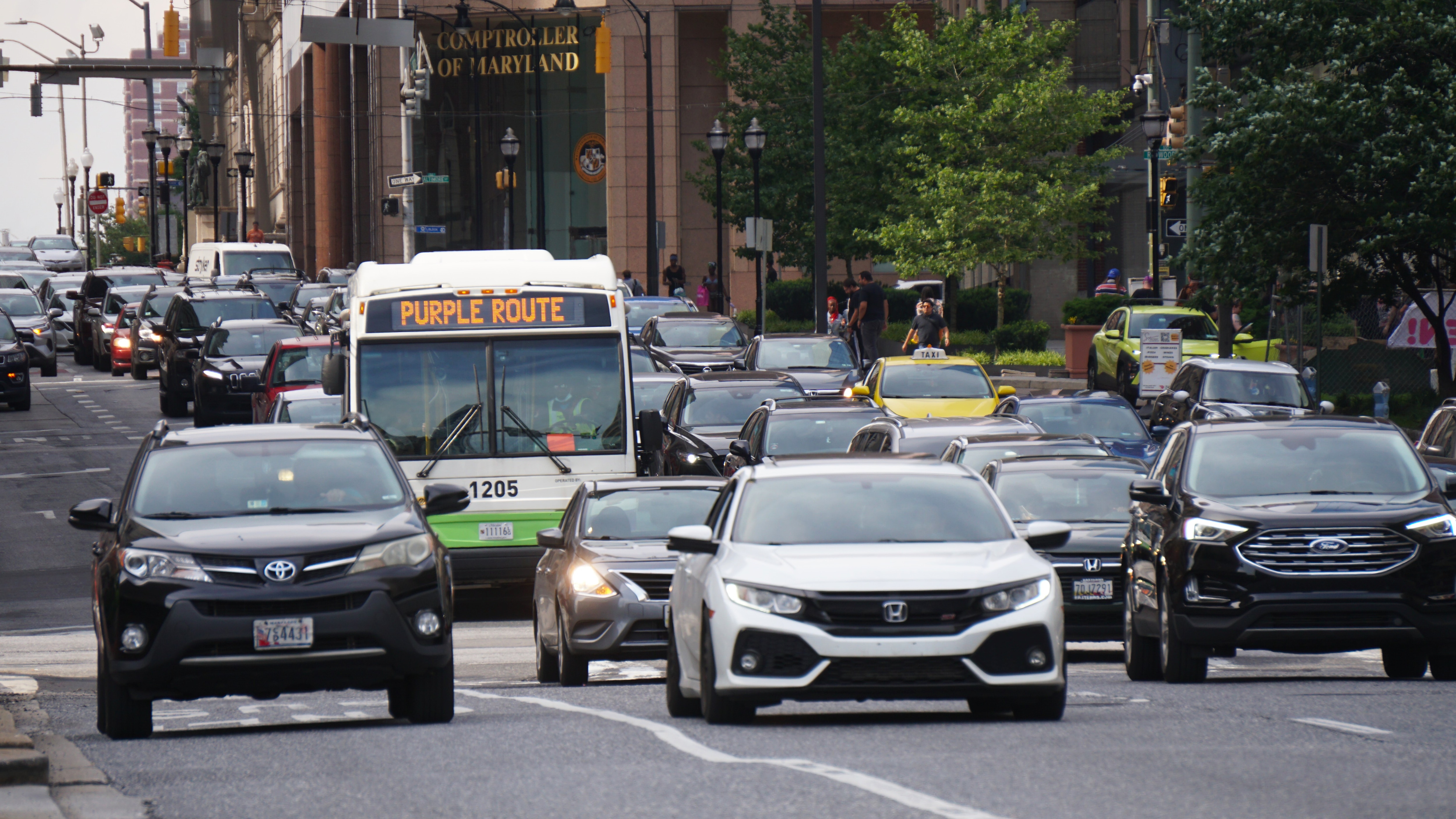 A white bus with a sign saying "PURPLE ROUTE" at the top is seen amongst a crowd of moving cars.