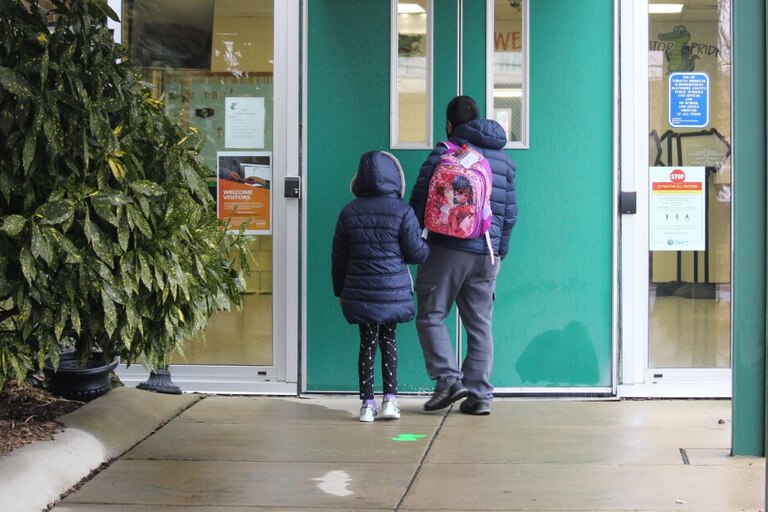 An adult walks a child to the front of a school building.