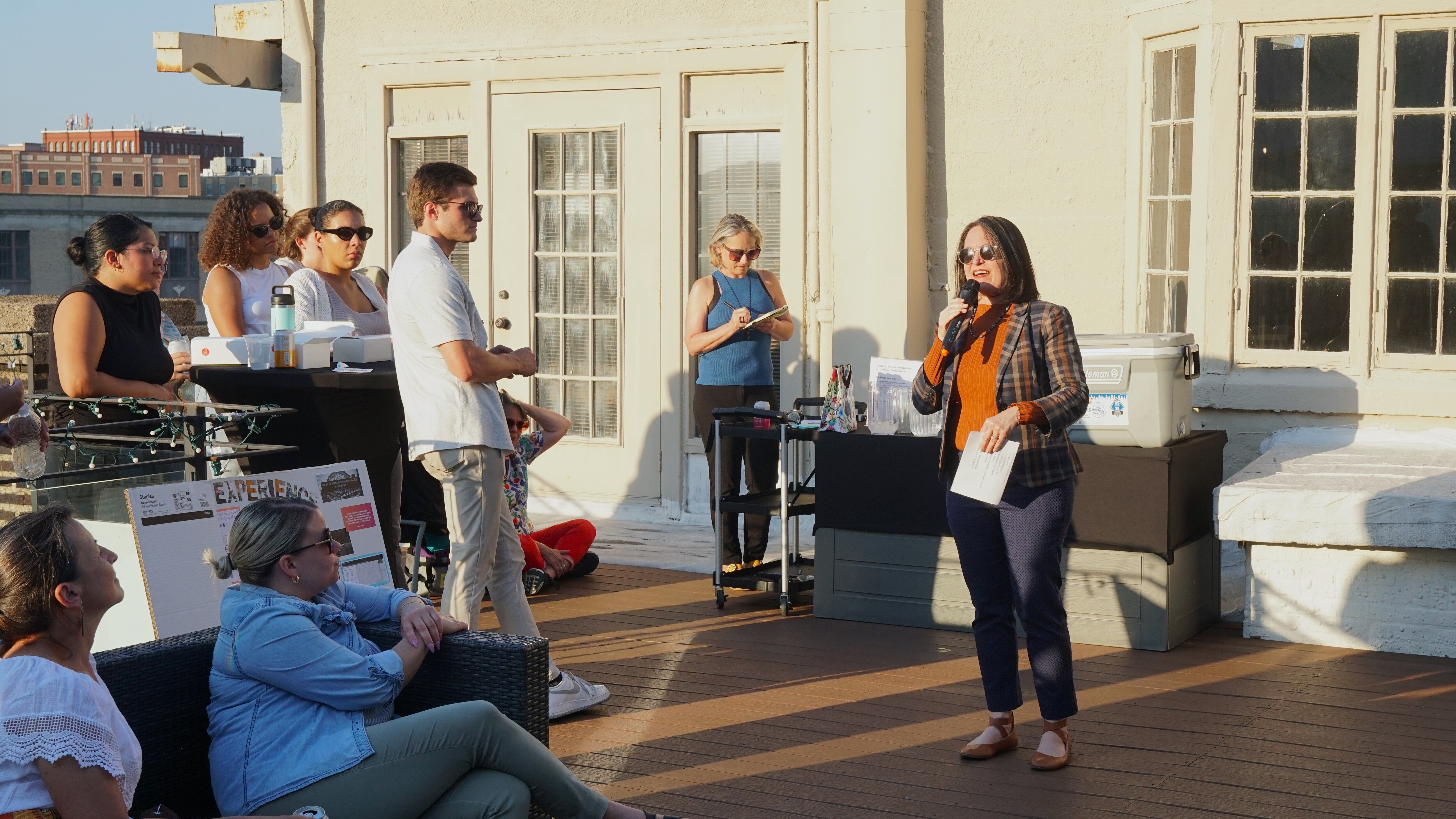A woman holding a microphone and a white piece of paper stands on a deck around a group of people and gives a speech.