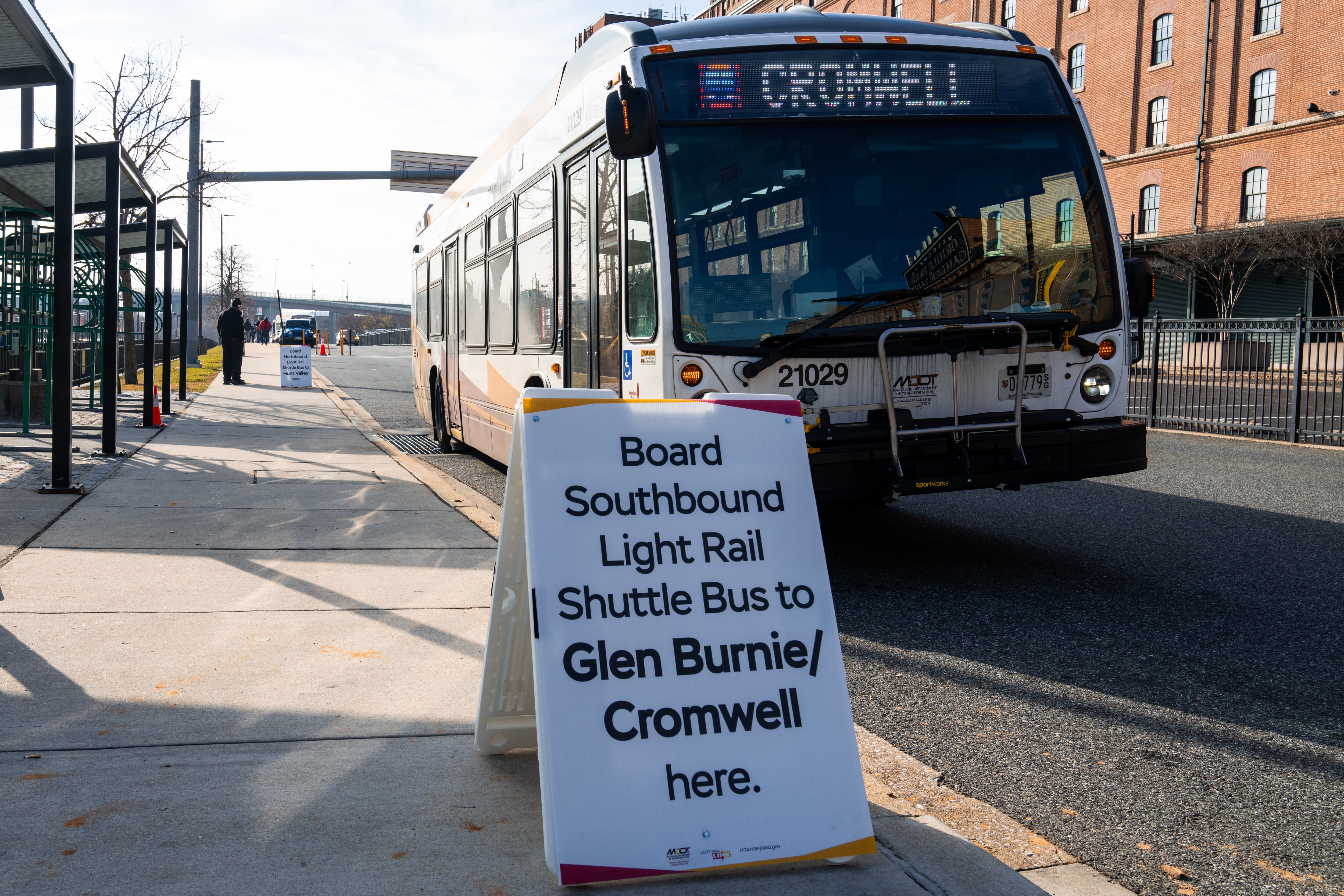 A southbound shuttle bus pulls up in front of a sign reading "Board Southbound Light Rail Shuttle Bus to Glen Burnie/Cromwell here."