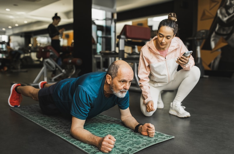 An aged man doing a plank with an assistant crouching next to him.