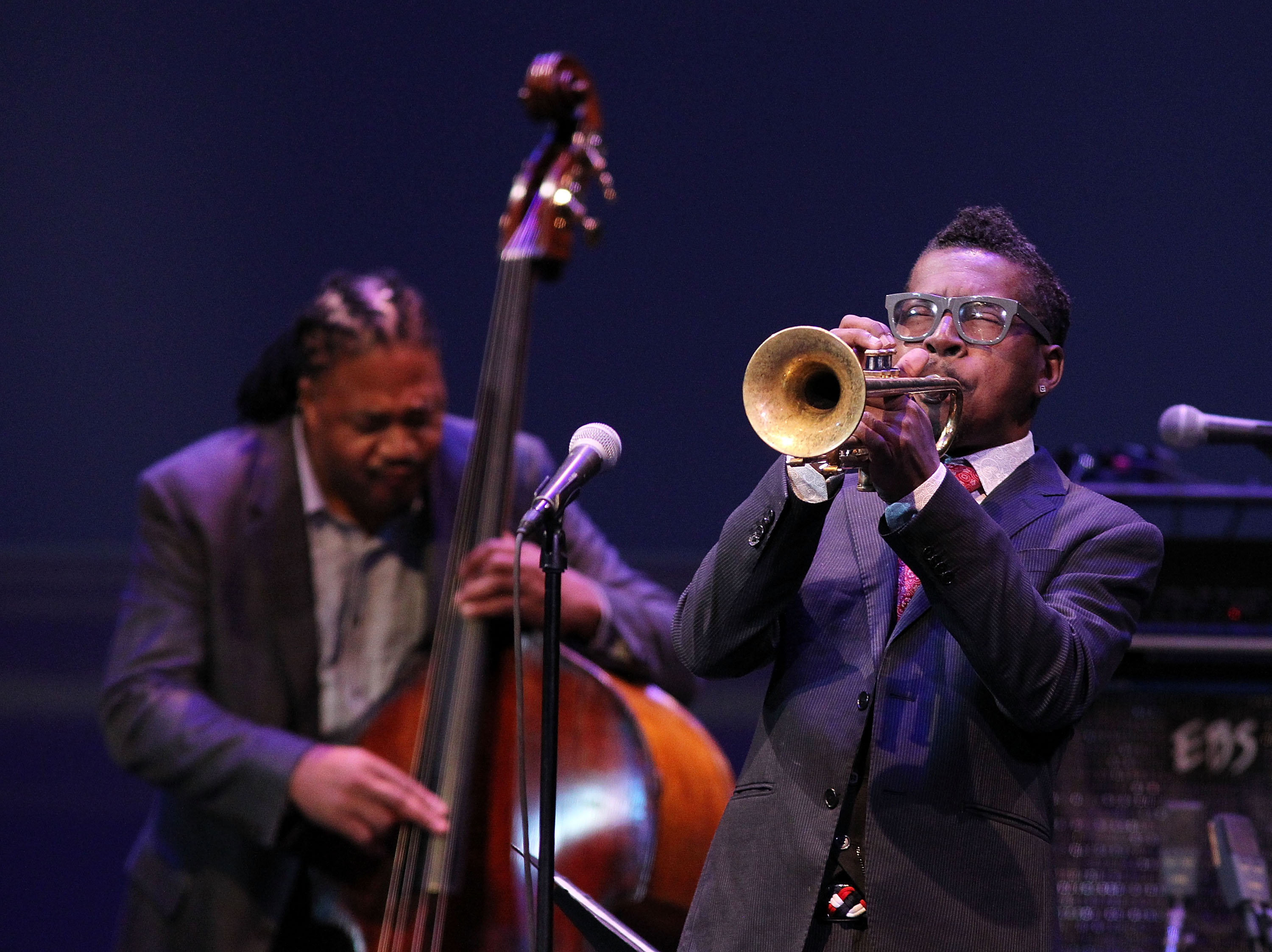 A man in a dark suit wearing glasses blows on trumpet with his eyes closed. Behind him another musician plucks a double bass.