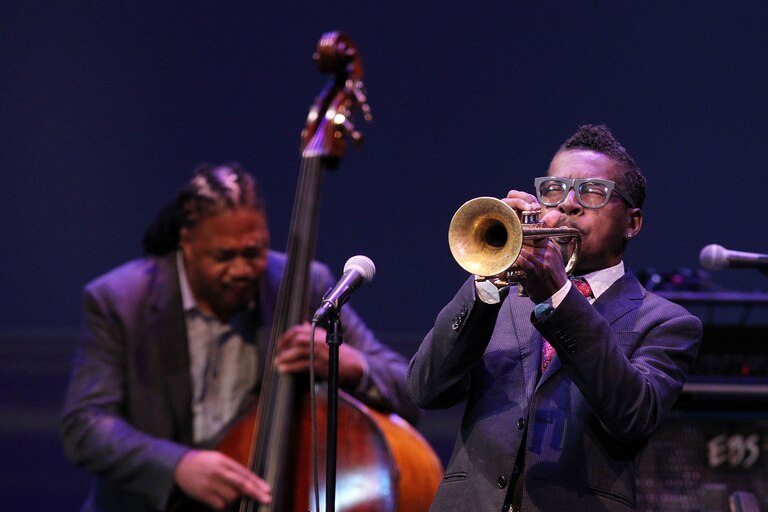 A man in a dark suit wearing glasses blows on trumpet with his eyes closed. Behind him another musician plucks a double bass.