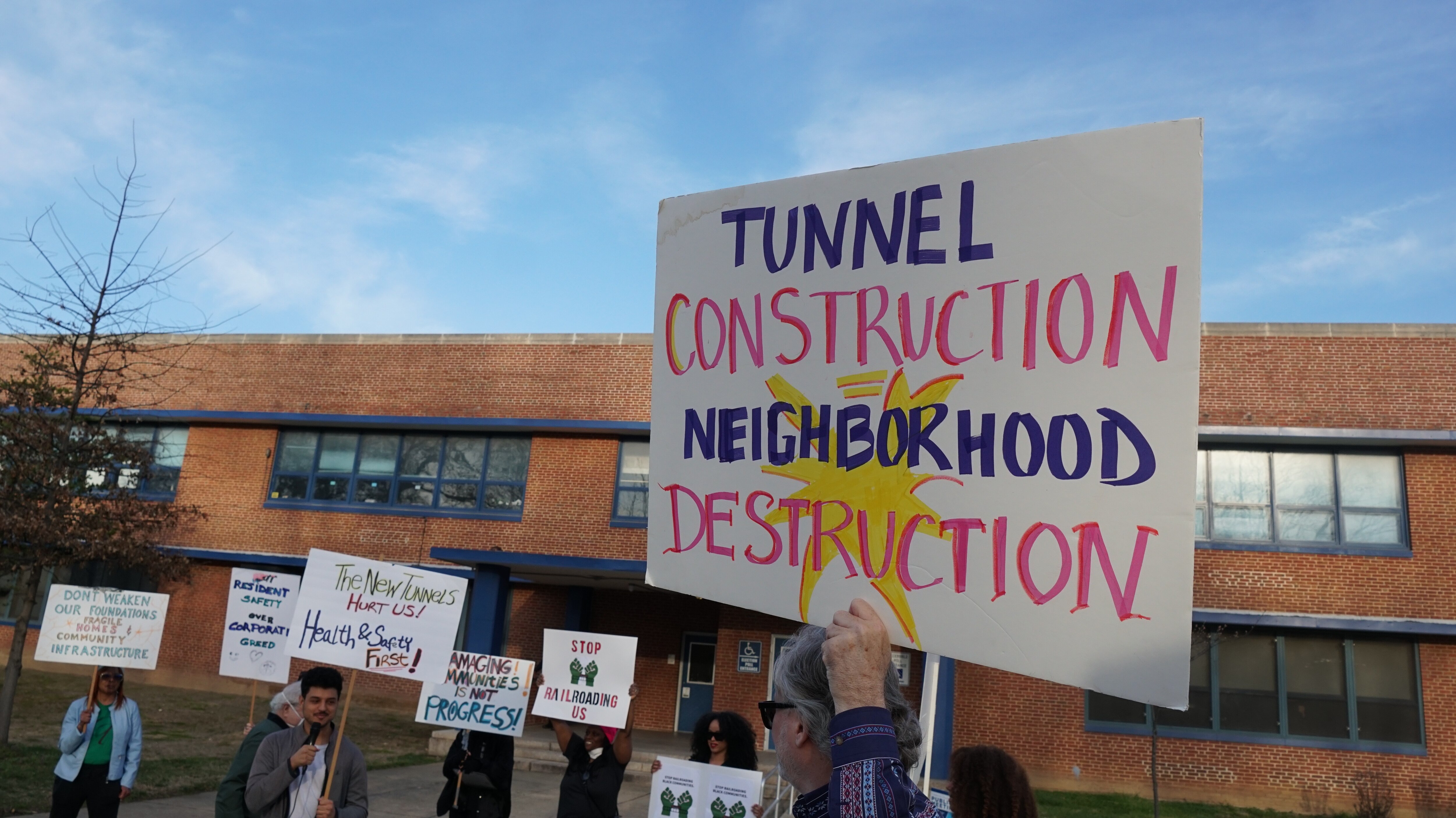 People hold a series of signs, including one that reads, 'TUNNEL CONSTRUCTION = NEIGHBORHOOD DESTRUCTION' in front of a brown school building.