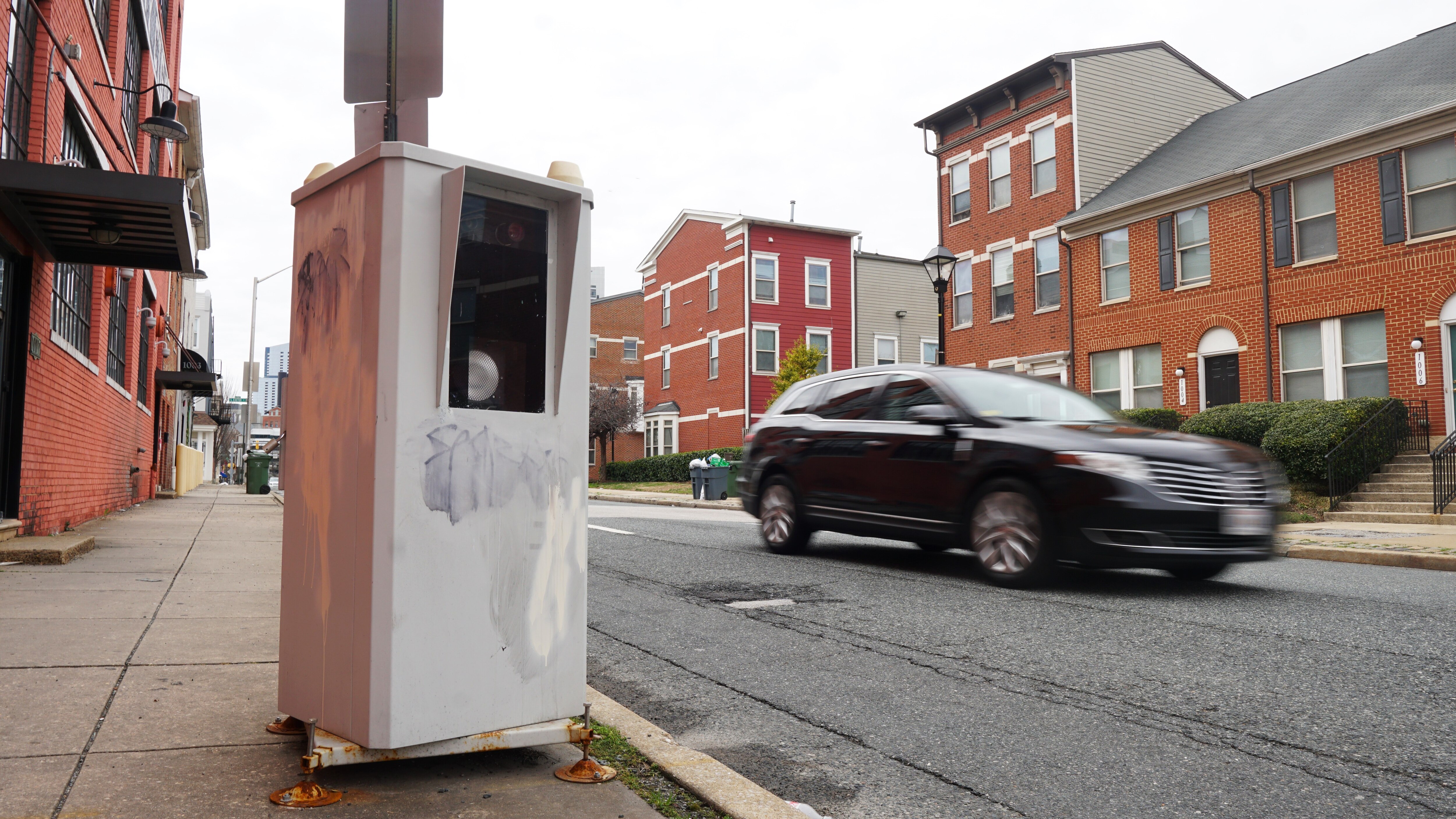 A rectangular grey box that houses an automated traffic camera stands on a city sidewalk as a blurred black SUV drives by.