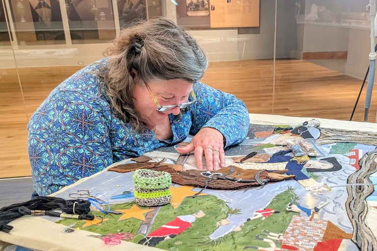Volunteer Nathalie Smith works on the Maryland panel of America's Tapestry. The size of the piece makes it difficult to reach under and push the needle up through the linen backing.