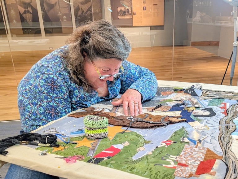 Volunteer Nathalie Smith works on the Maryland panel of America's Tapestry. The size of the piece makes it difficult to reach under and push the needle up through the linen backing.
