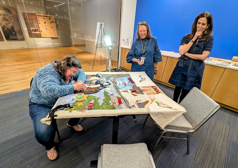 Nathalie Smith works on the Maryland panel of America's Tapestry at the Maryland History and Culture Center as curator Catherine Rogers Arthur, center, and CEO Katie Caljean watch.