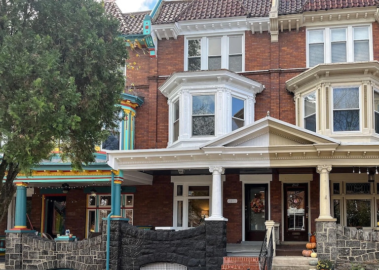A historic red brick row house sits between two other homes. It has a raised porch and white molding and finishes. The house to the left of it has bright blue, yellow and red finishings. The home to it's right has beige finishing.