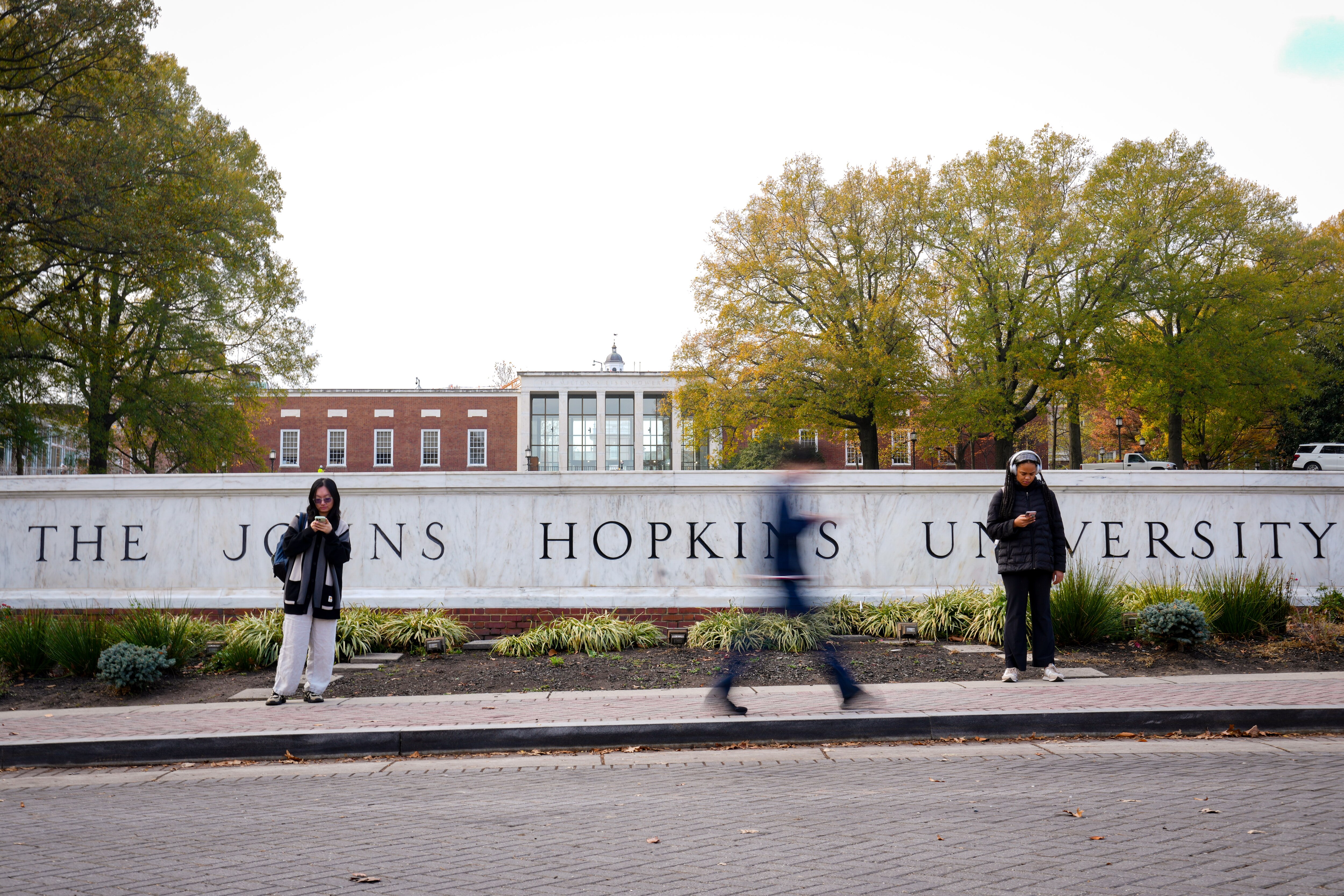 Some students wait for a shuttle while others walk past the Johns Hopkins University sign welcoming people to the Homewood Campus from Charles Street in Baltimore, Md. on Wednesday, November 20, 2024.