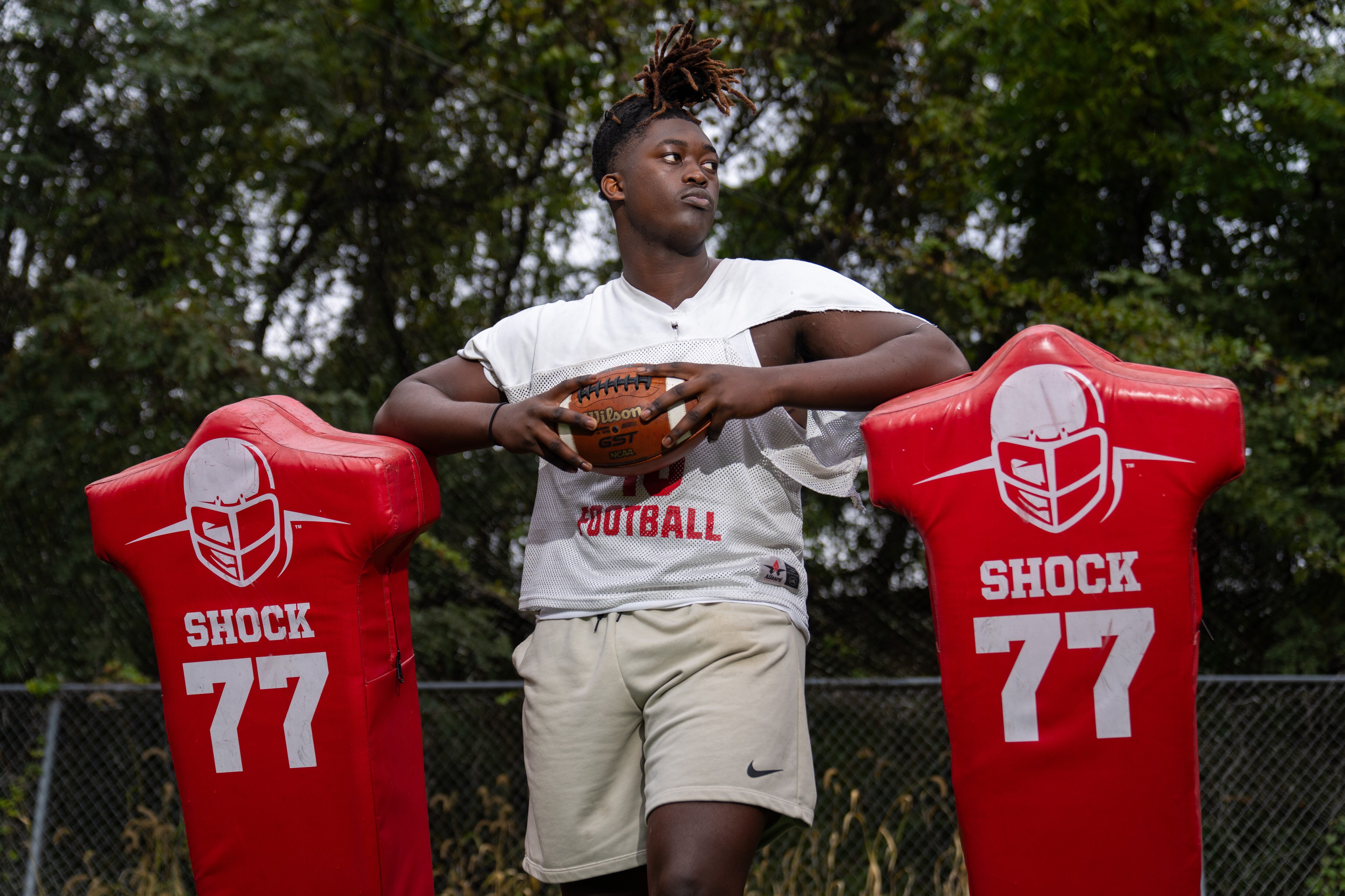 Ernest Willor looks away from the camera for a portrait in front of a red blocking sled while holding a football.