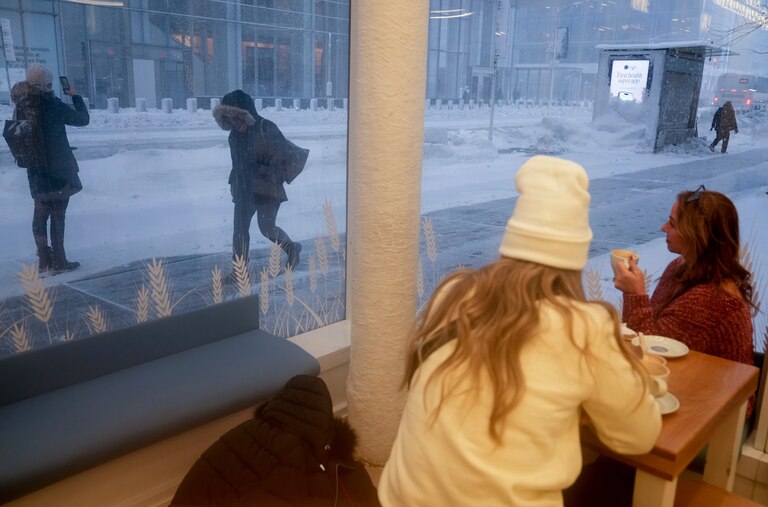 Women sipping coffee at a coffee shop watch as pedestrians pass during a snow storm, Monday, Feb. 23, 2026, in New York.