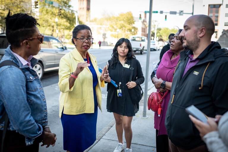 A group of adults stand around talking on the sidewalk of a downtown street on a sunny day.