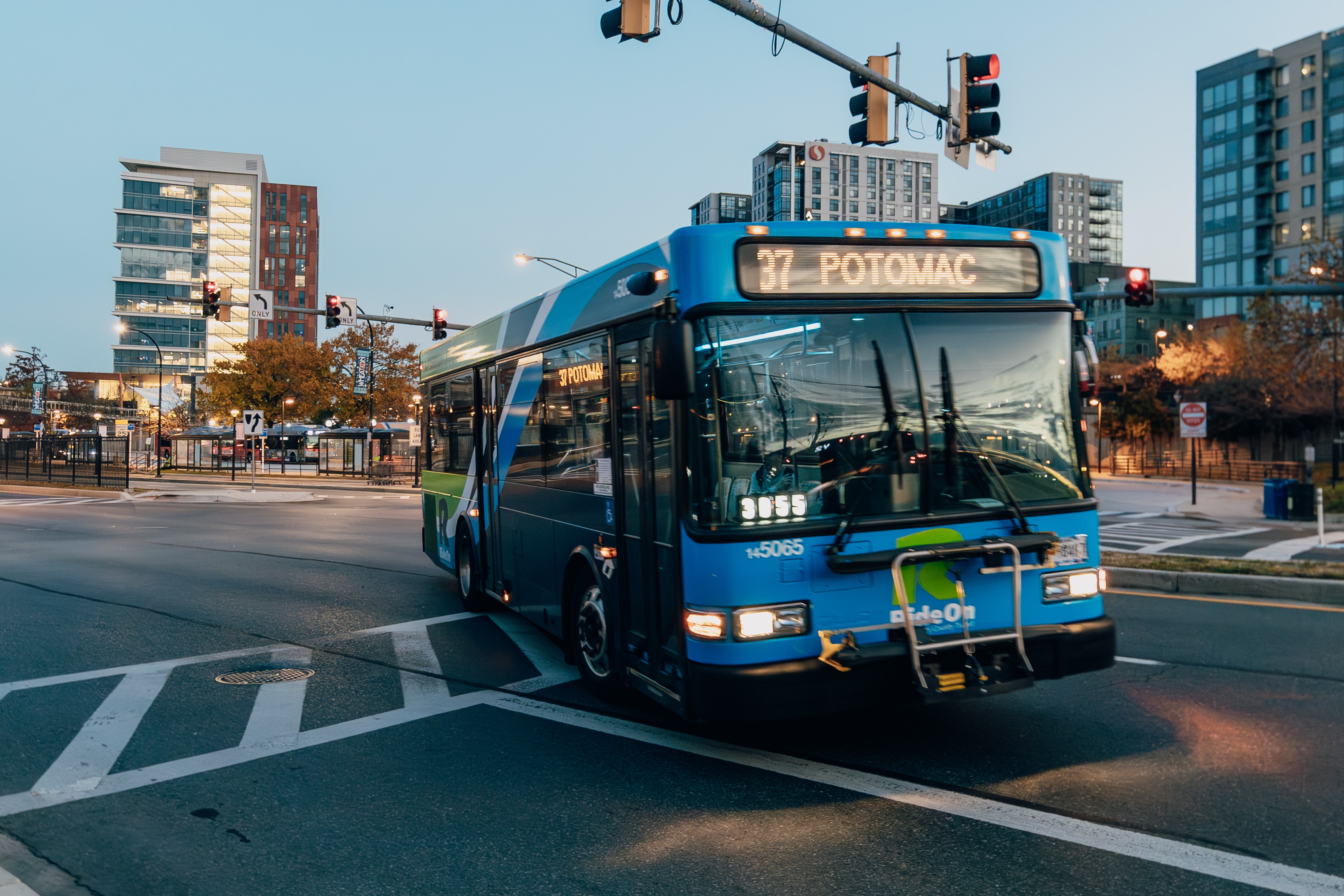 A blue bus makes a turn at a city intersection at dusk. The bus has a digital sign at the top that reads "37 POTOMAC"
