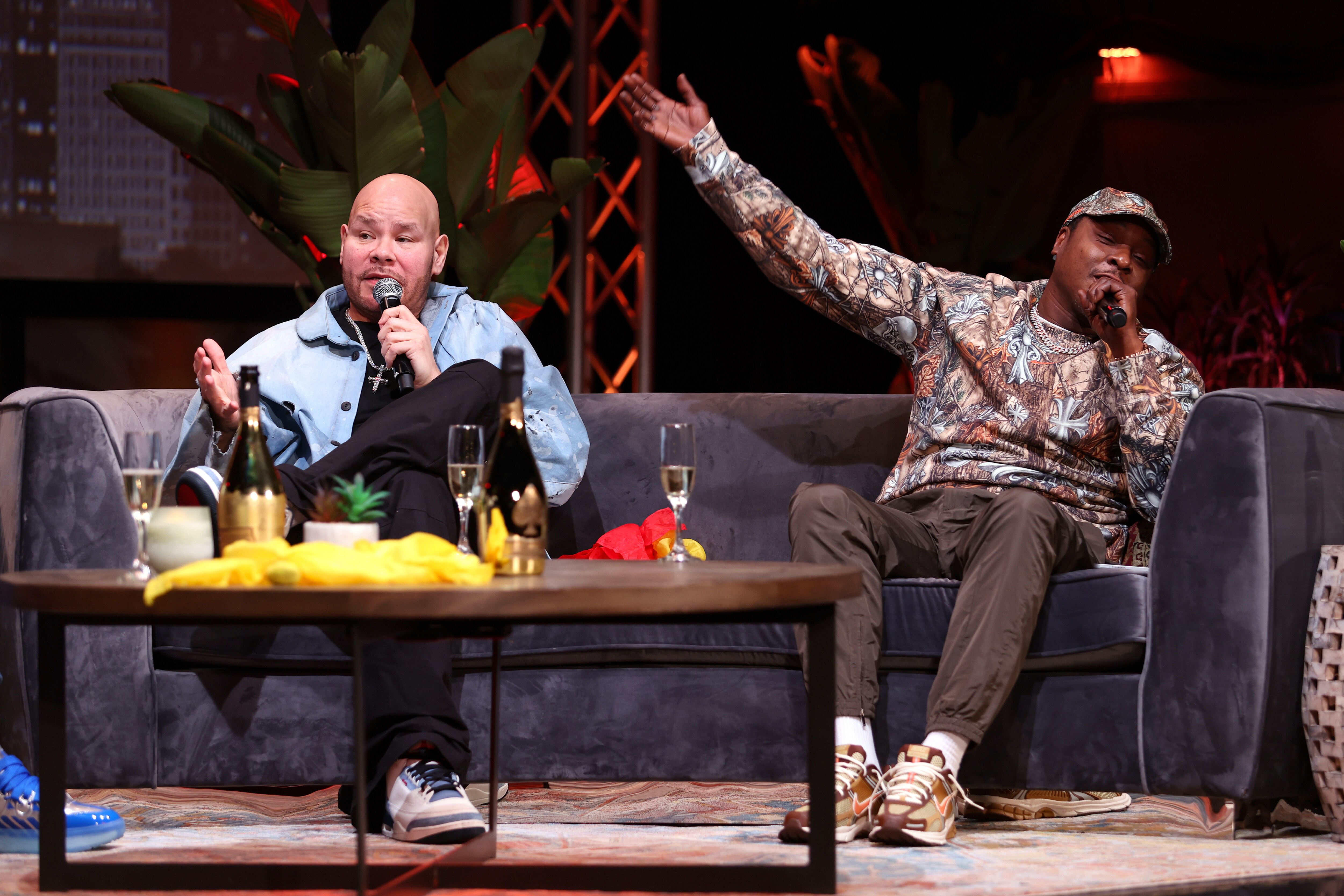 Two men sit on a couch holding microphones. The man on the right holds his right arm up in the air. A coffee table in front of them has champagne flutes and a bottle of champagne.