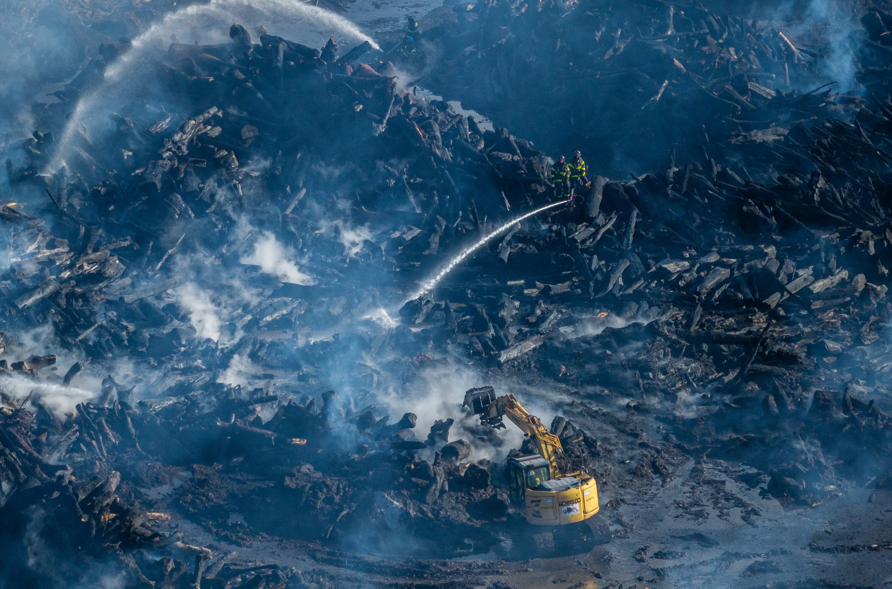 Firefighters spray water on the smoldering wood piles as an excavator moves logs at the Camp Small city wood recycling facility Friday morning.