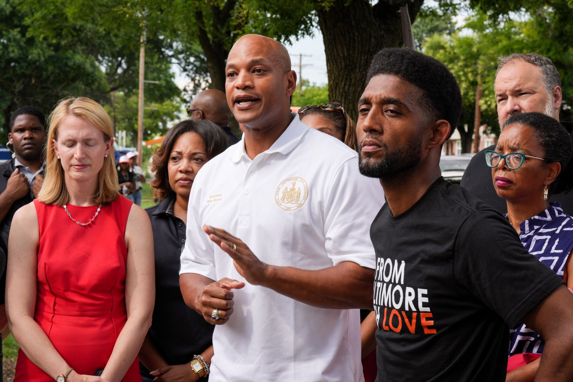 Maryland Gov. Wes Moore, center, speaks with reporters outside a community center in Brooklyn on July 4, 2023, to discuss the mass shooting that took place nearby a few days earlier, standing alongside Baltimore Mayor Brandon Scott and other elected Maryland and local government officials. (Ulysses Muñoz / The Baltimore Banner)