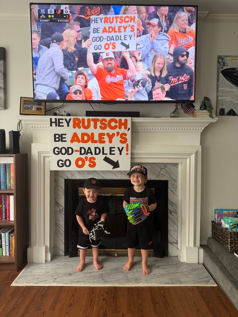 Josiah and Casey Dells' two sons, Adley and Brooks, pose with a still shot of young Adley at an Orioles game. Josiah Dells holds a sign asking Adley Rutschman to be the "God-Dadley"