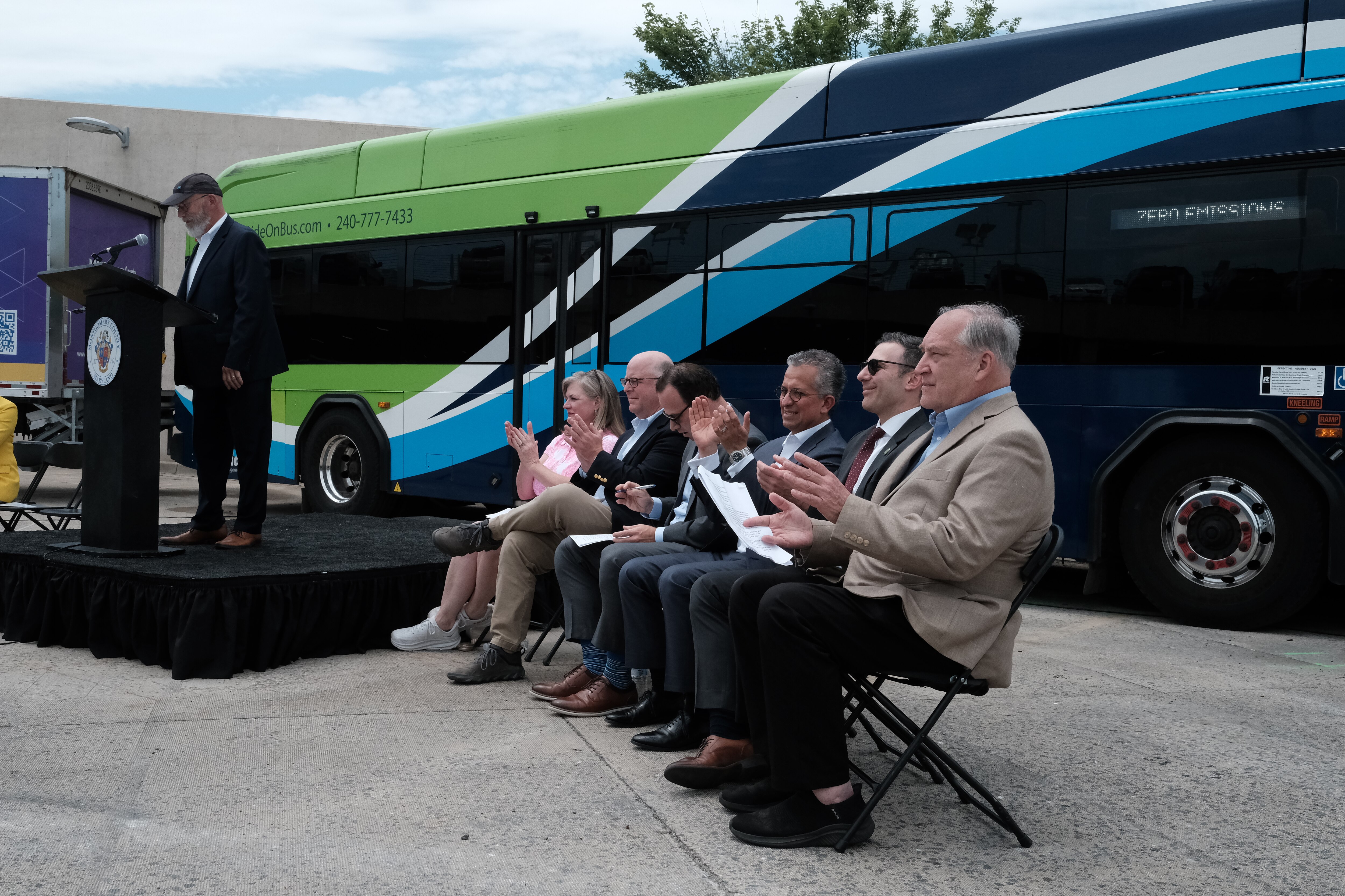 Seven people sit in a line of chairs in front of a large green and blue bus next to a stage on which a man is speaking at a podium.