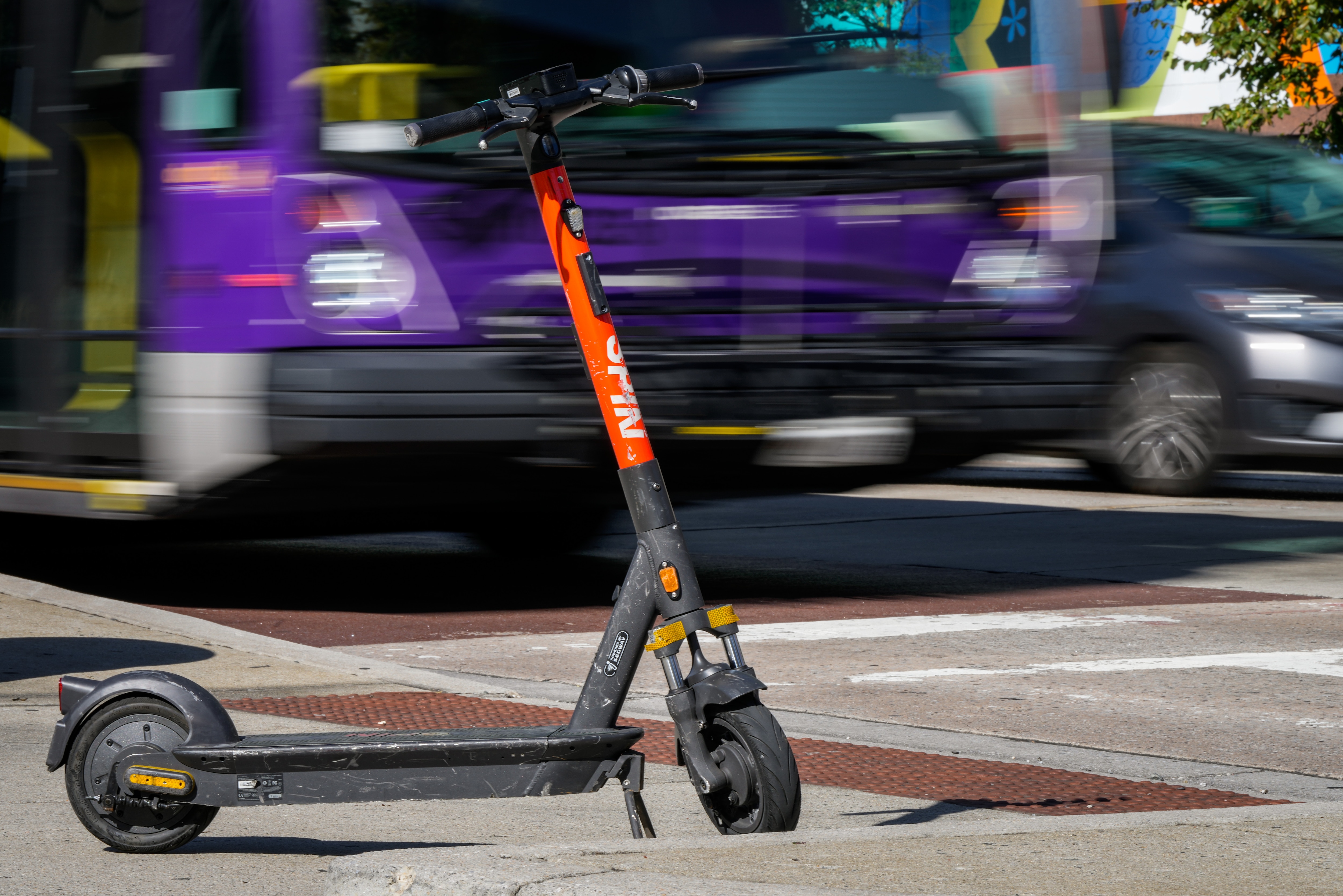 A purple bus is captured in slow shutter speed going by a parked orange Spin scooter.