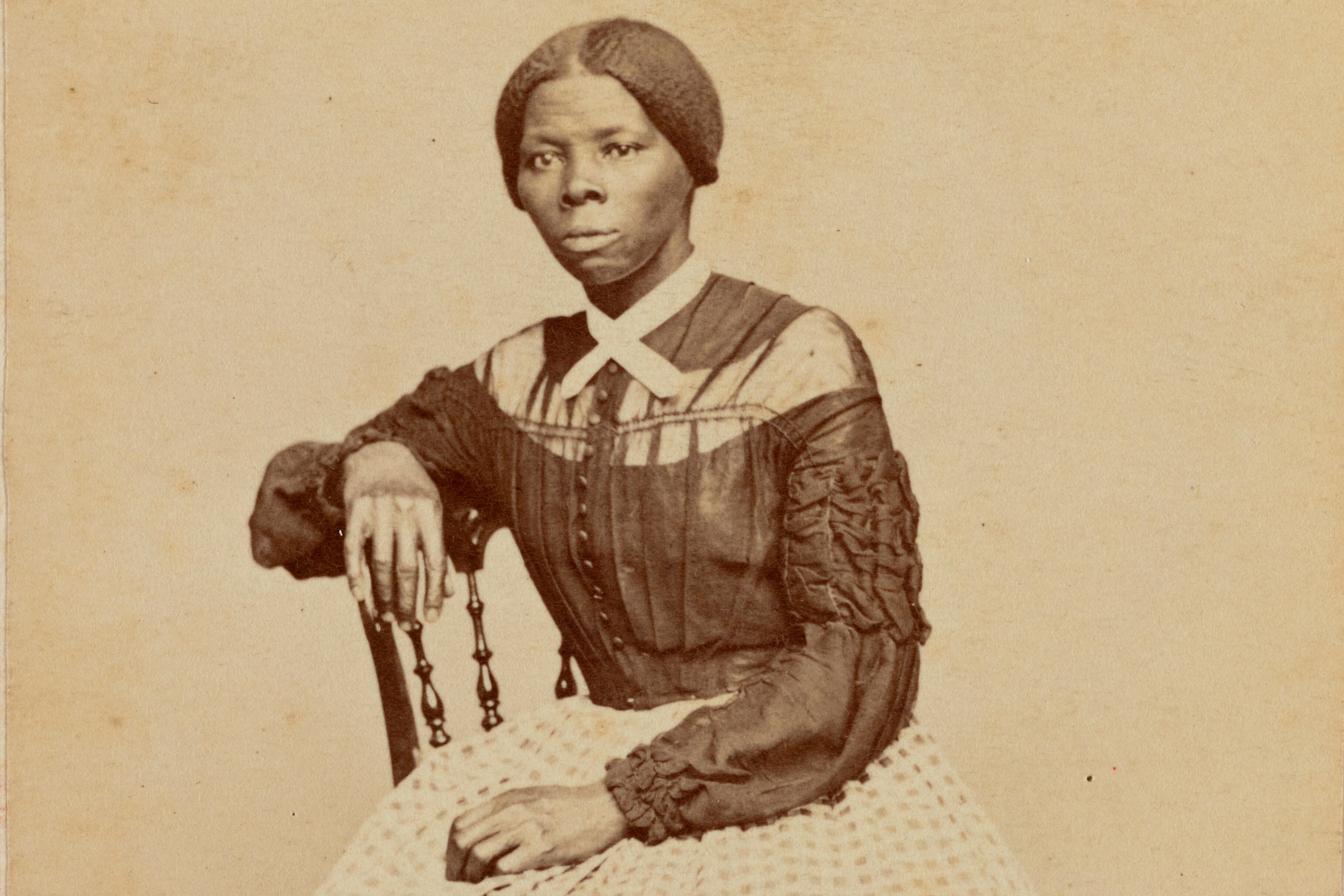 Photograph shows Harriet Tubman (1822-1913) at midlife. She is seated, turned toward the left. One hand rests on the back of a wooden chair, another rests in her lap.